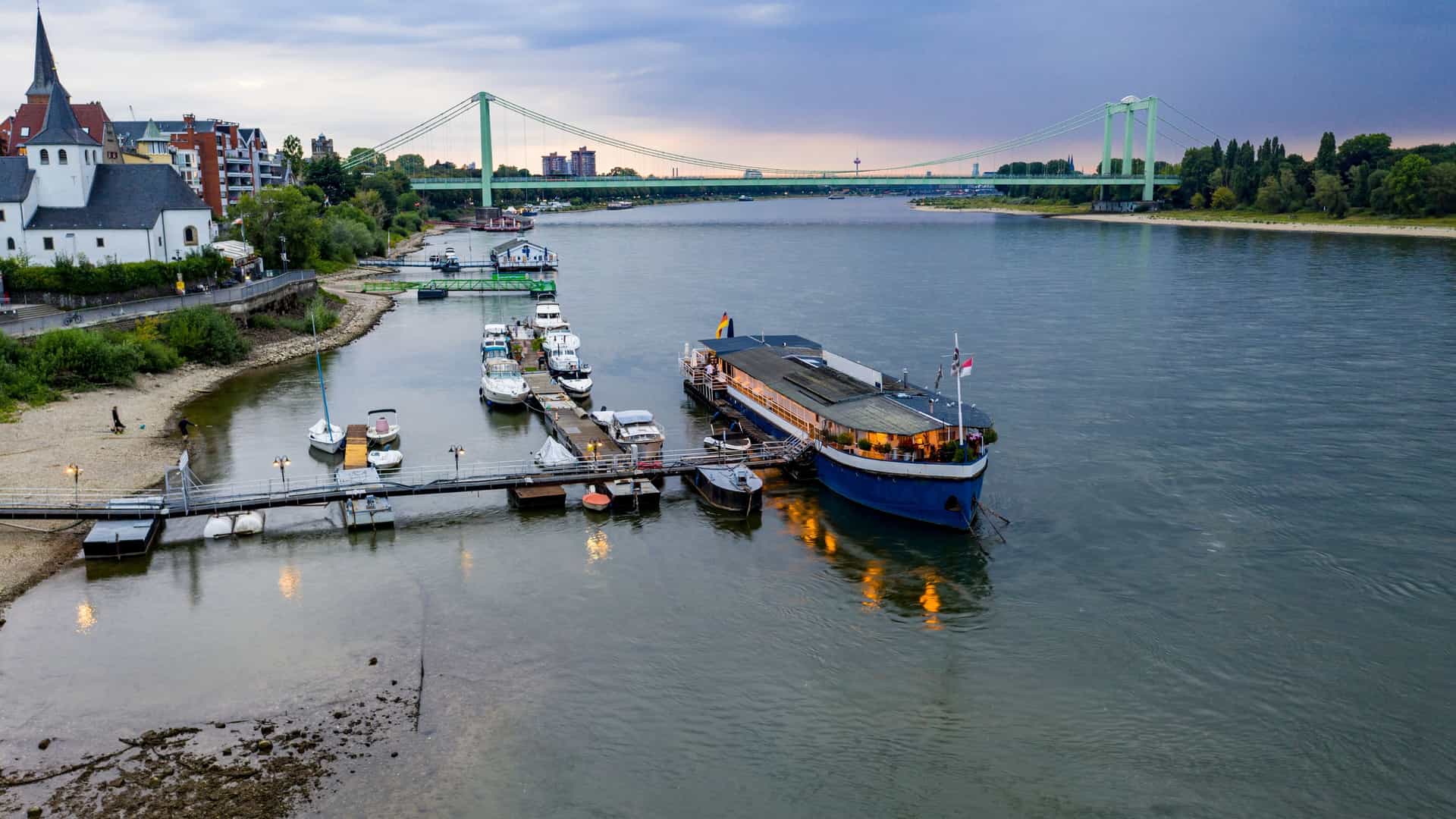  A permanently moored boat on the Rhine River has been converted into a unique restaurant, featuring a multi deck structure with an expansive outdoor dining terrace on the upper level. The image captures the vessel's rustic appearance and its prime riverside location, with the dark river water and a small barge visible in the background.