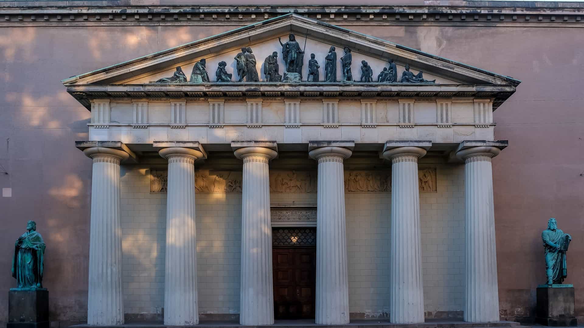  The grand Neoclassical facade of Copenhagen Cathedral (Vor Frue Kirke) is defined by a deep portico with six massive Doric columns supporting a triangular pediment. The entrance is flanked by two large bronze statues, depicting Moses and King David.
