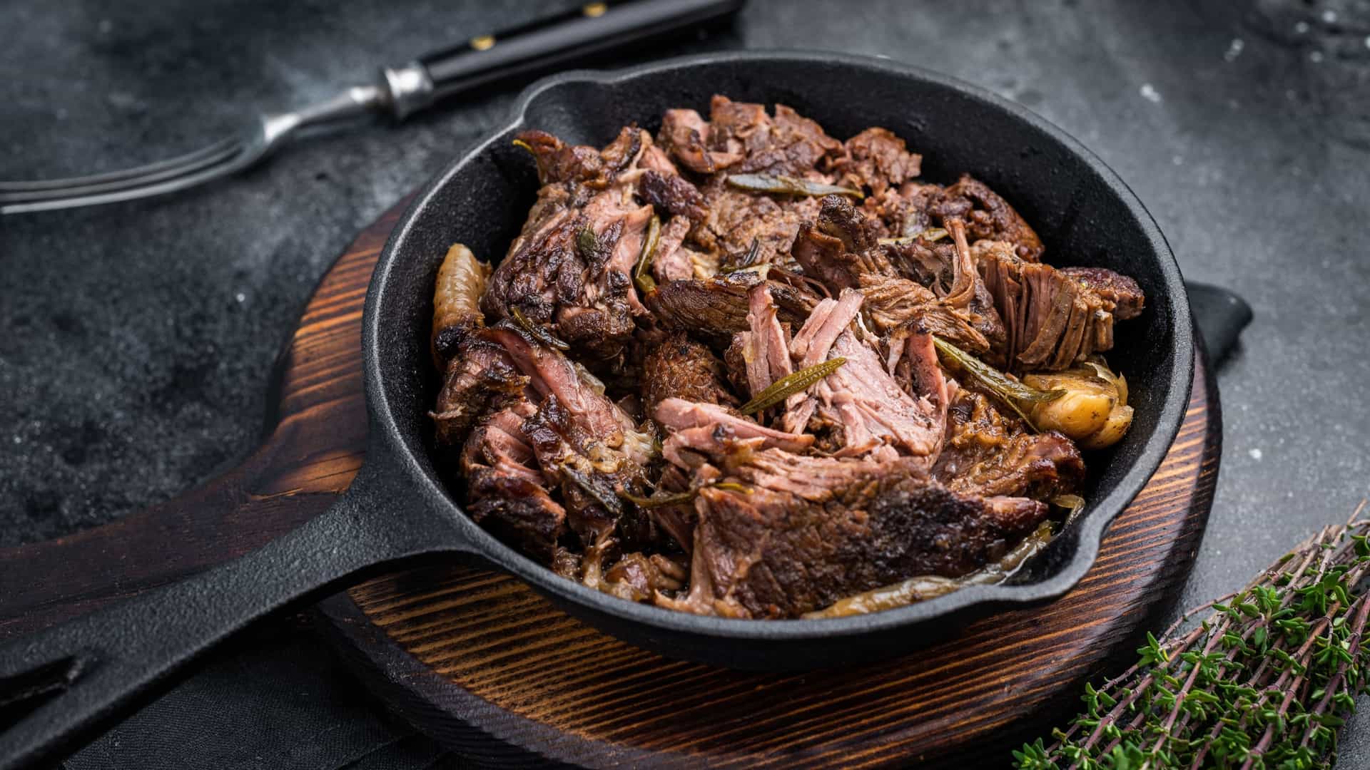  A top down, close up view of a Beef Bourguignon stew served in a black pan, set against a black background. The stew is rich and dark, featuring tender pieces of beef, whole pearl onions, and sliced carrots, garnished with fresh herbs.