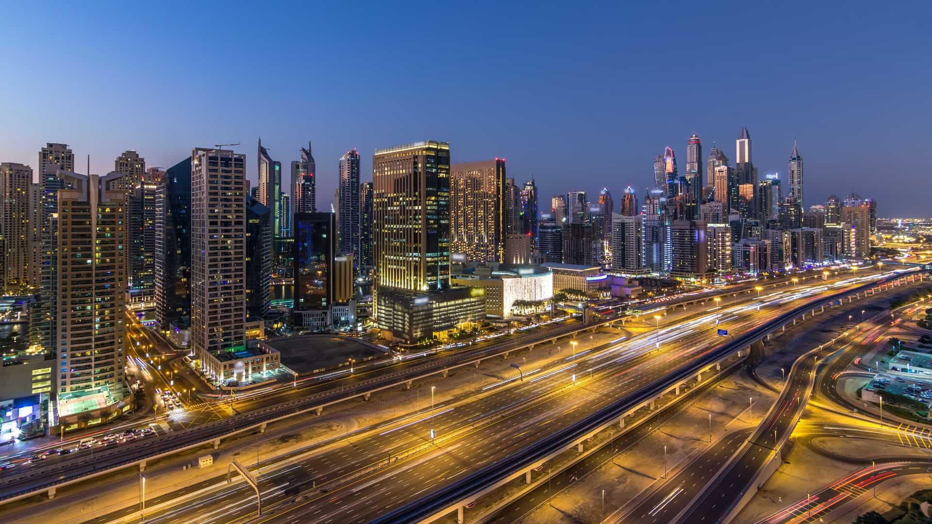  An aerial or high angle view of a multi lane highway, Sheikh Zayed Road, in Dubai. The road is busy with traffic, and is flanked by a dense cluster of modern skyscrapers that make up the Dubai Marina skyline. The scene is captured at either dusk or night, with the streetlights and building lights illuminating the area.