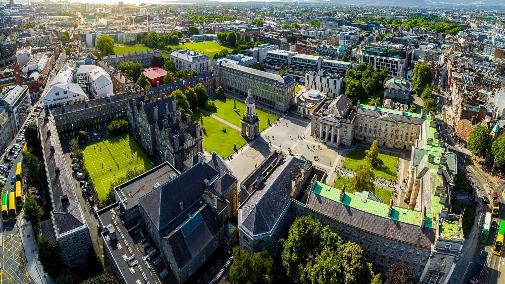  An aerial view of Trinity College Dublin, showing the large, historic campus quadrangle, central green spaces, and surrounding classical architecture, including the famous Campanile (bell tower) and various institutional buildings, all within the urban setting of Dublin, Ireland.