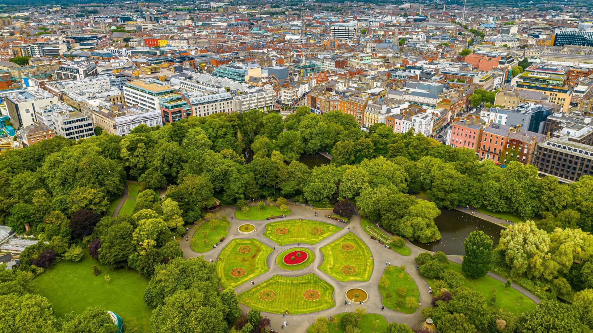  An aerial or drone view of St. Stephen's Green, a large rectangular public park in the center of Dublin, Ireland. The image shows the park's lush green grass, walking paths, ornamental lake, and various trees, which may show autumn colors. The park is surrounded by city streets and buildings, showcasing its location as a green oasis within the urban landscape.