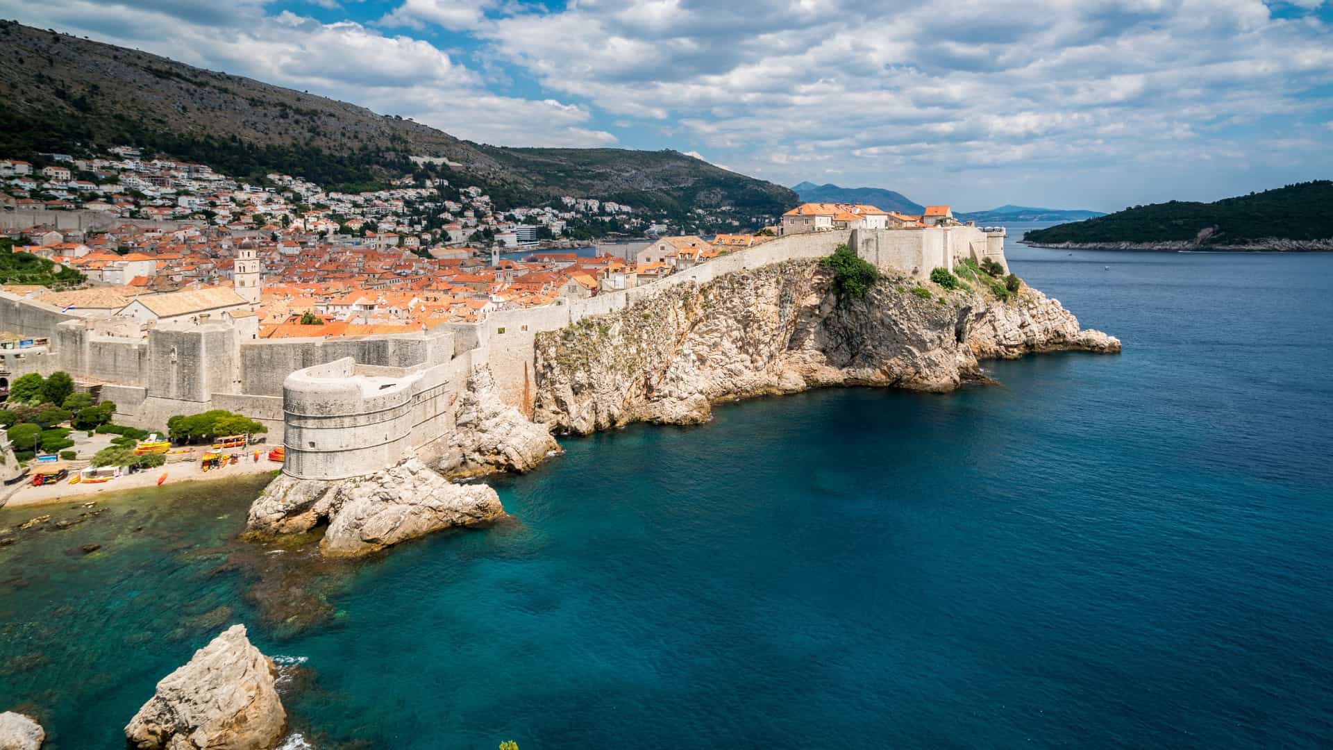  high angle, panoramic view of the massive stone city walls of Dubrovnik, Croatia, jutting out over the bright turquoise blue waters of the Adriatic Sea. The old town's terracotta roofed buildings are visible behind the protective medieval fortifications, with steep, rocky hills rising in the background under a cloudy sky.