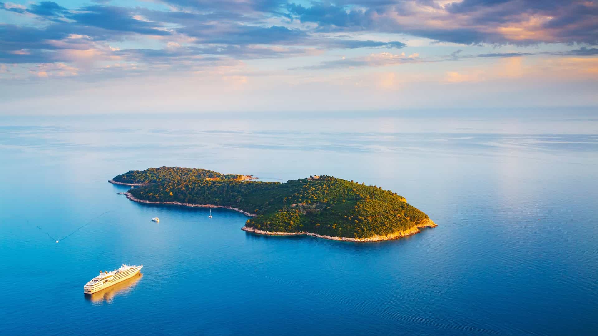   A scenic view of Lokrum Island, showing a small, tranquil salt lake known as the "Dead Sea" surrounded by dense green vegetation, including lush trees and rocks. The calm, clear water of the lake reflects the natural surroundings.