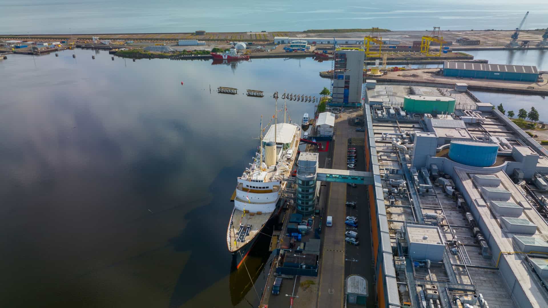  A port side view of the Royal Yacht Britannia, the former royal yacht of the British monarchy, moored in a harbor. The white hull and masts of the vessel are prominent, and the British flag is visible. The yacht is docked alongside a pier, with a building in the background.