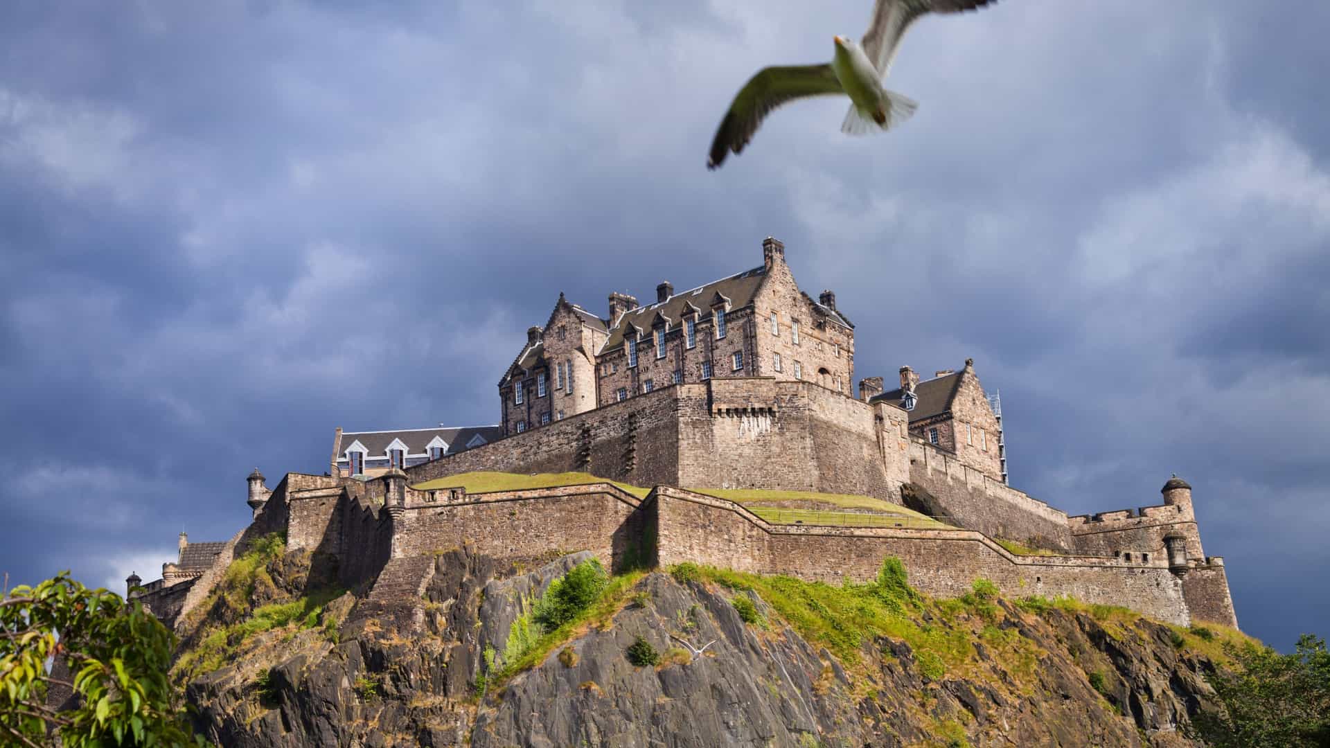  A scenic view of Edinburgh Castle perched majestically on Castle Rock, a large volcanic crag that rises above the surrounding city. The historic fortress, with its prominent stone walls and towers, is shown from a distance, highlighting its commanding position and rocky foundation.