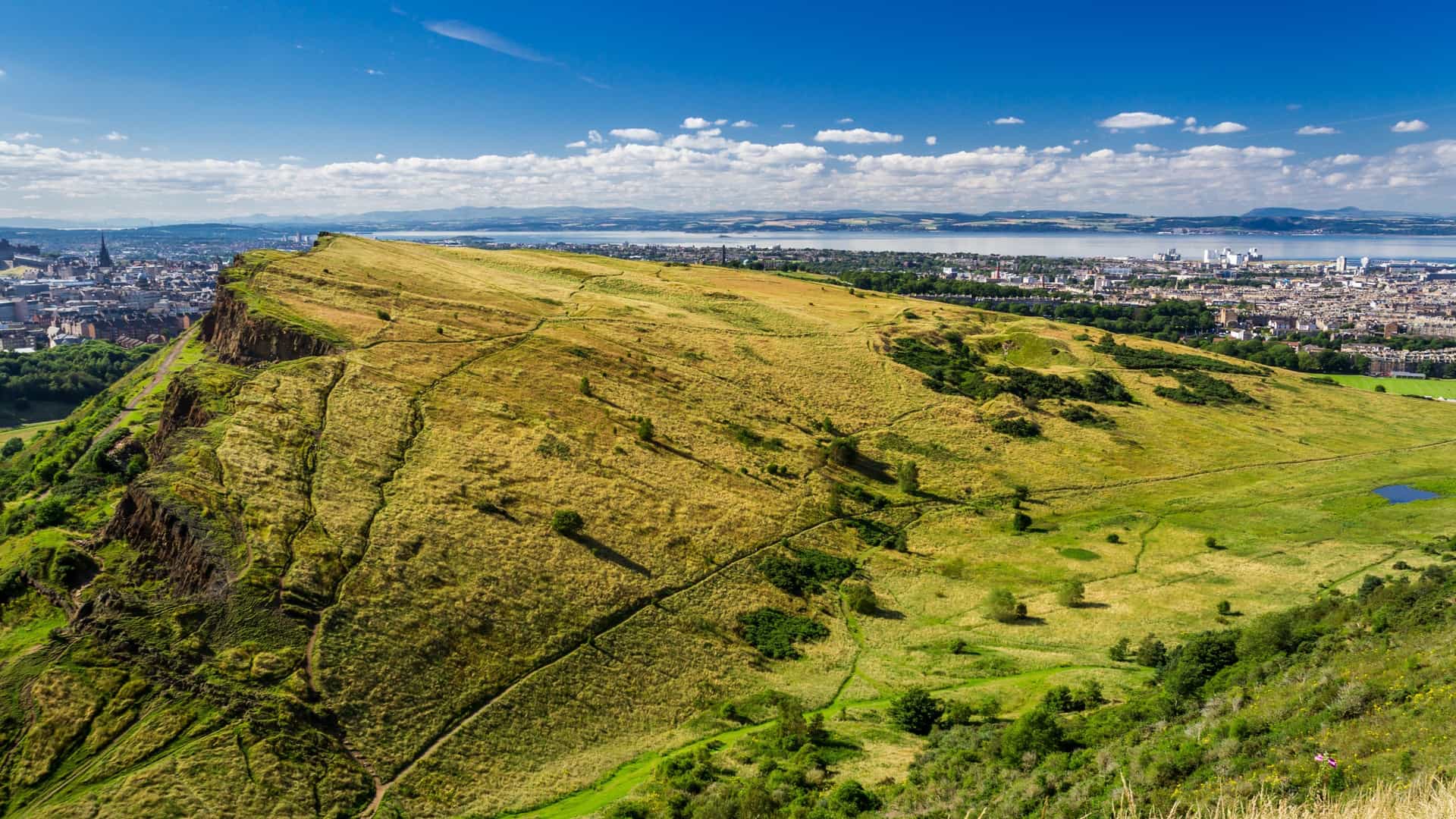 An elevated view of the Edinburgh cityscape from the top of Arthur's Seat. The city's buildings, including the castle and other prominent landmarks, are visible, stretching out towards the sea. The landscape is a mix of urban development and the green hills of Holyrood Park.