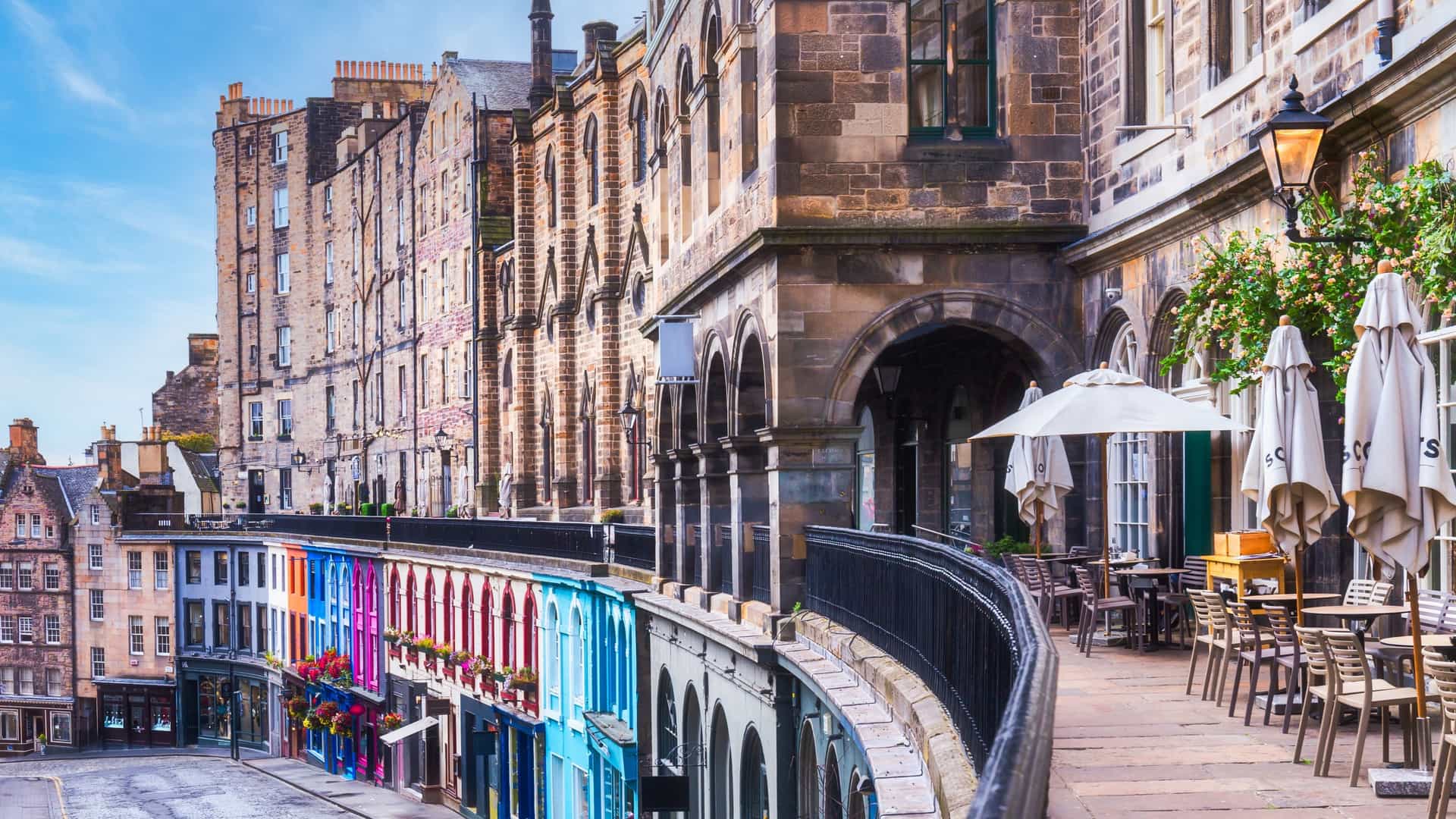 A captivating view of the curved, sloping Victoria Street in Edinburgh's Old Town. The scene is dominated by the street's famous colorful facades—a mix of historic stone buildings painted in vibrant hues. The street is paved with cobblestones and is lined with eclectic shops and businesses, contributing to the street's popular, historic, and lively atmosphere, which is often associated with famous fictional locations.