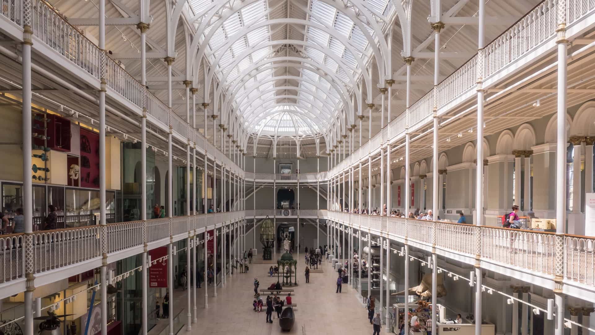 A spectacular interior view of the Grand Gallery at the National Museum of Scotland in Edinburgh. The photo captures the towering Victorian cast-iron and glass atrium, which is flooded with natural light from the massive skylight roof. The gallery features multiple tiered balconies and a dramatic, vertical display called the 'Window on the World', showcasing a diverse collection of objects and surrounded by the museum's historic, ornate architecture.