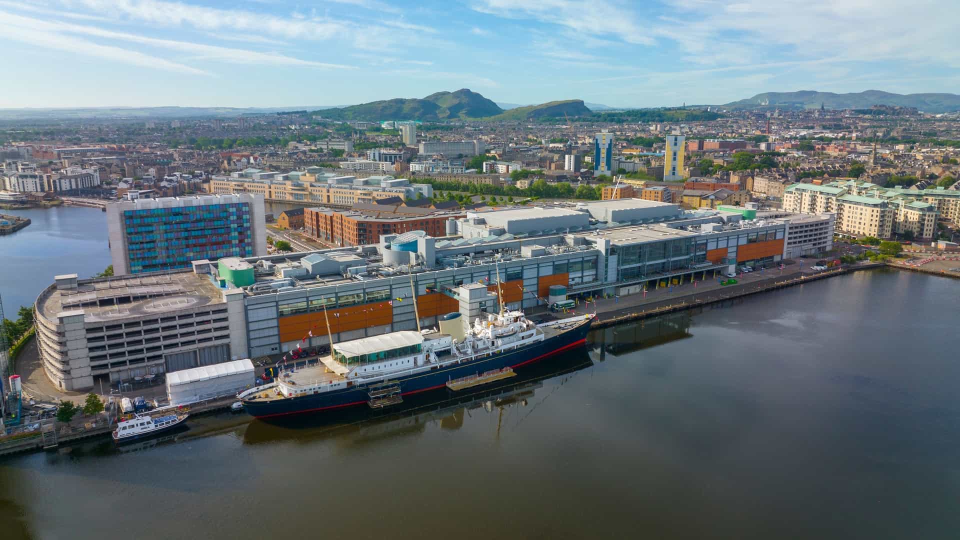  A view of the Royal Yacht Britannia, the former royal yacht of the British monarchy, now a museum ship moored at the Port of Leith in Edinburgh, Scotland. The image shows the elegant vessel with its all white hull and prominent red trim, set against a calm body of water. The ship's deck and masts are visible, and the surrounding dock area and buildings of the port can be seen in the background, including a large building on the left with a unique architectural design.