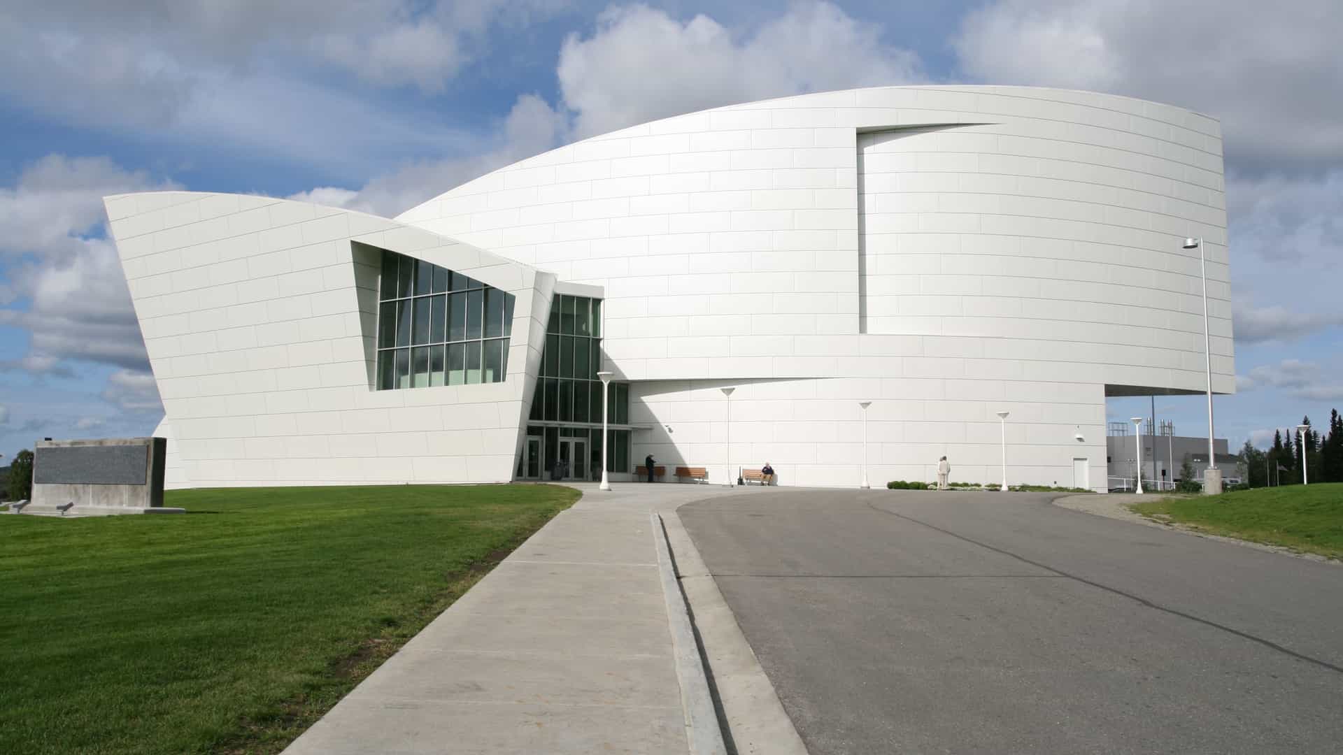 A modernist building with stark, white, angular architecture against a light-colored sky, identifying it as the University of Alaska Museum of the North in Fairbanks. The building's design is sculptural, meant to evoke Alaskan forms like glaciers or alpine ridges, featuring a sweeping, curved, and cantilevered upper section that contrasts with its lower part, with large windows visible on the facade.