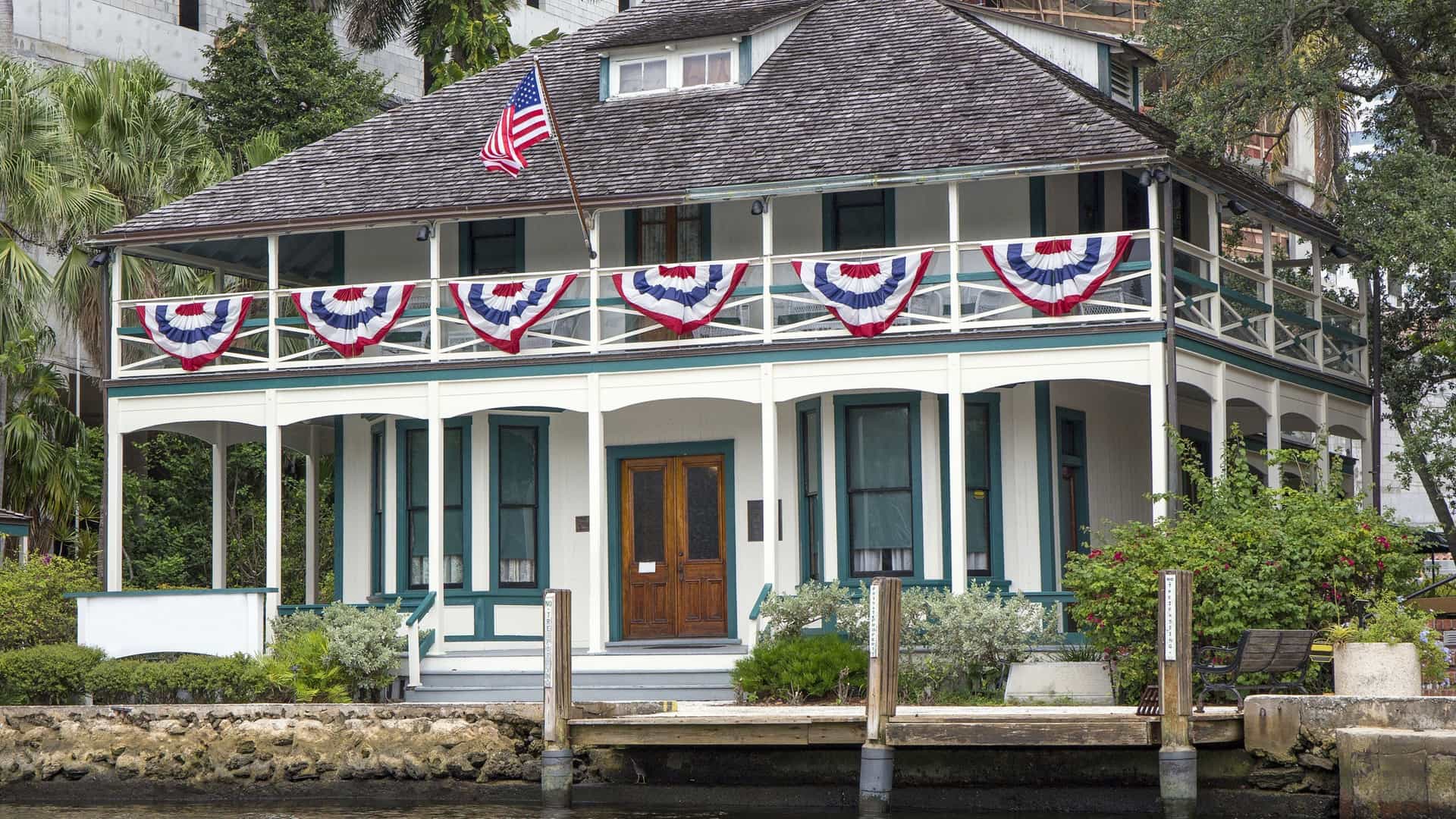 A daytime photograph of the Historic Stranahan House Museum in Fort Lauderdale, Florida. This two-story, white, wood-frame building, which is the oldest surviving structure in Broward County, features wide wrap-around porches on both levels. The house is situated on the bank of the New River, with a lush lawn and tropical vegetation visible in the foreground, and modern high-rise buildings of the Fort Lauderdale skyline visible in the background across the river.