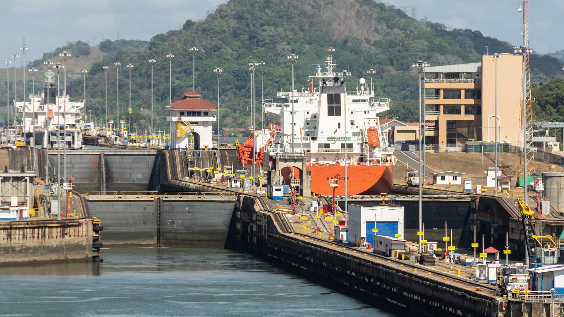  An aerial view of the Miraflores Locks on the Panama Canal. A large ship is visible within one of the lock chambers, surrounded by the concrete walls and gates of the lock system. The image captures the scale and engineering of the canal, with the surrounding landscape and visitor center also in view.