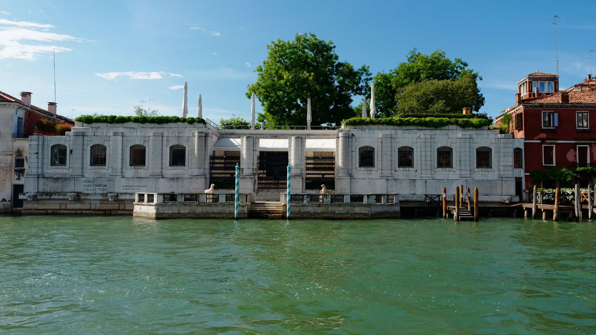 A long, low-rise, classic white stone palazzo, the Palazzo Venier dei Leoni, is visible on the edge of the Grand Canal in Venice, Italy. This building houses the Peggy Guggenheim Collection. It features a flat roof with greenery, a central water entrance, and a row of arched windows. The structure is set against a bright blue sky, with a smaller, traditional Venetian house visible to the right.