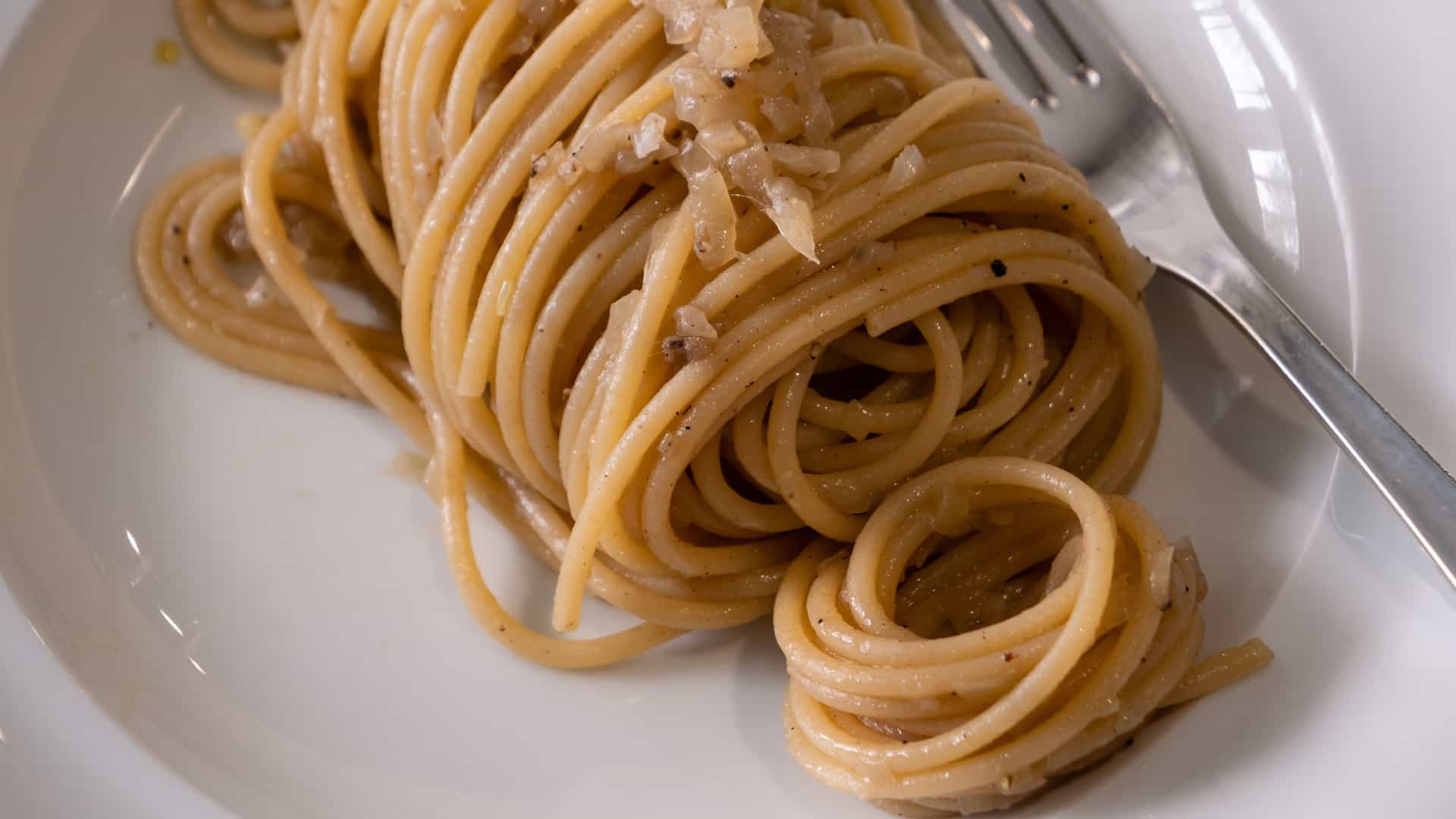 A close-up view of a plate of Bigoli in Salsa, a traditional Italian pasta dish from the Veneto region. The thick, whole-wheat Bigoli pasta is coated in a dark, savory sauce made from anchovies (or sardines) and slowly cooked white onions. The dish is typically garnished with chopped parsley and cracked black pepper.