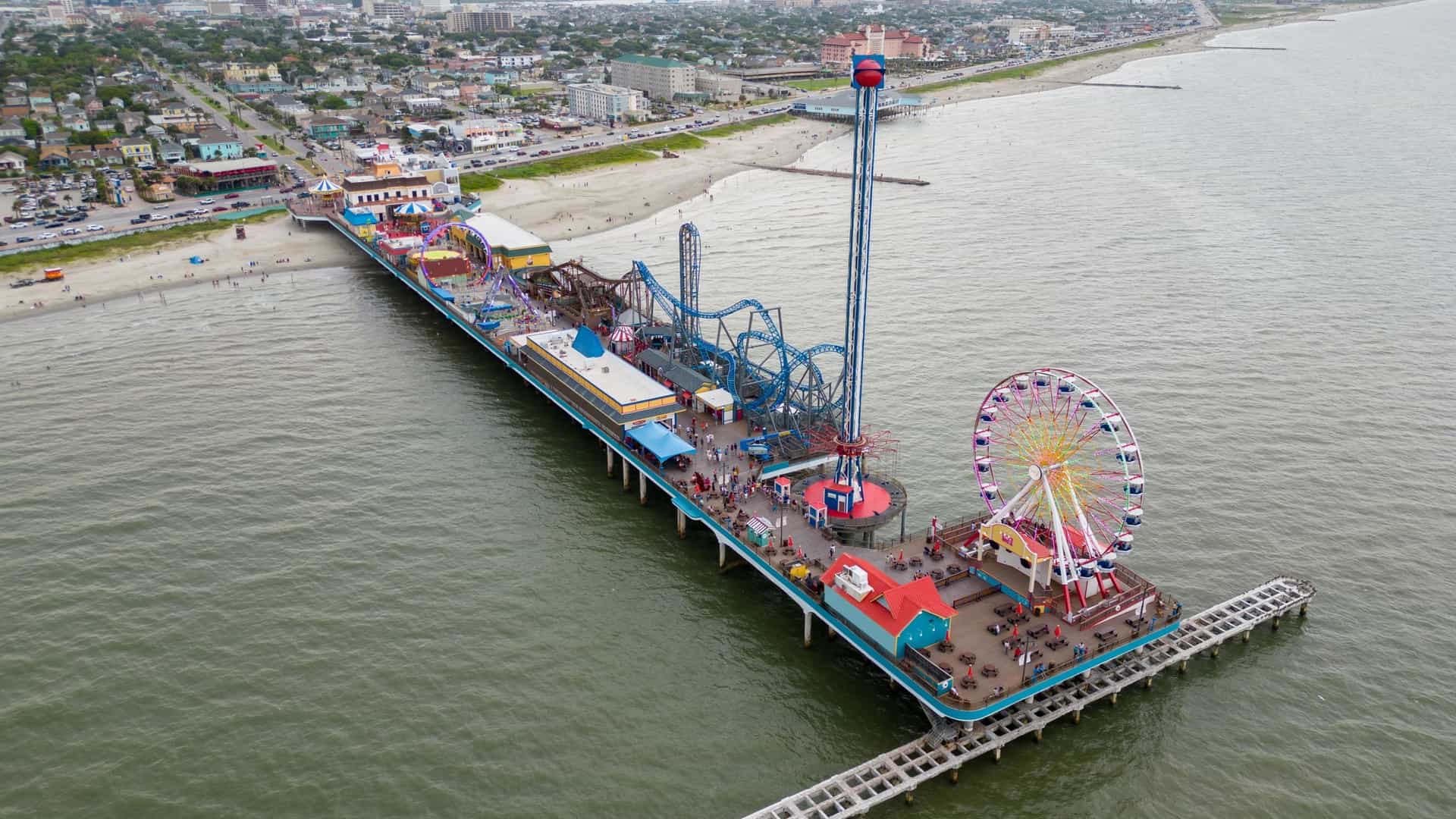 An aerial drone photo shows the historic Galveston Island Pleasure Pier and the surrounding beachfront. The pier, with its rides and attractions, extends out into the Gulf of Mexico, with the city's coastal buildings and seawall visible in the background.