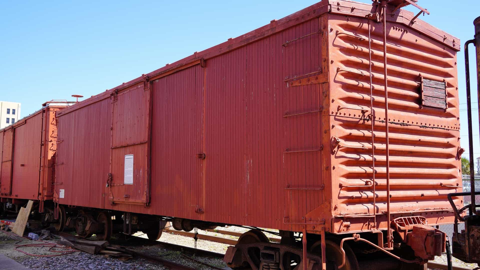A bright, low-angle photo of a faded red, antique freight boxcar on a railroad track at the Galveston Railroad Museum. The corrugated metal siding and ladder on the end of the boxcar show signs of age and weathering, set against a clear blue sky.