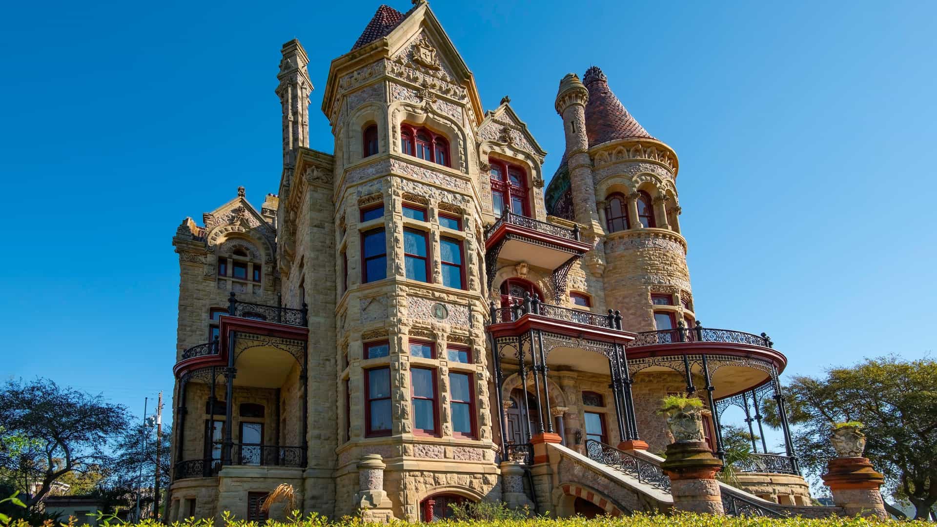 A low-angle view of the Bishop's Palace (Gresham's Castle) in Galveston, Texas, a grand Victorian-era mansion in the Châteauesque style. The façade is constructed of light-colored, intricately carved stone and features prominent turrets, pointed gables, and curved wrought-iron balconies. The building stands above a bright green lawn against a clear blue sky.