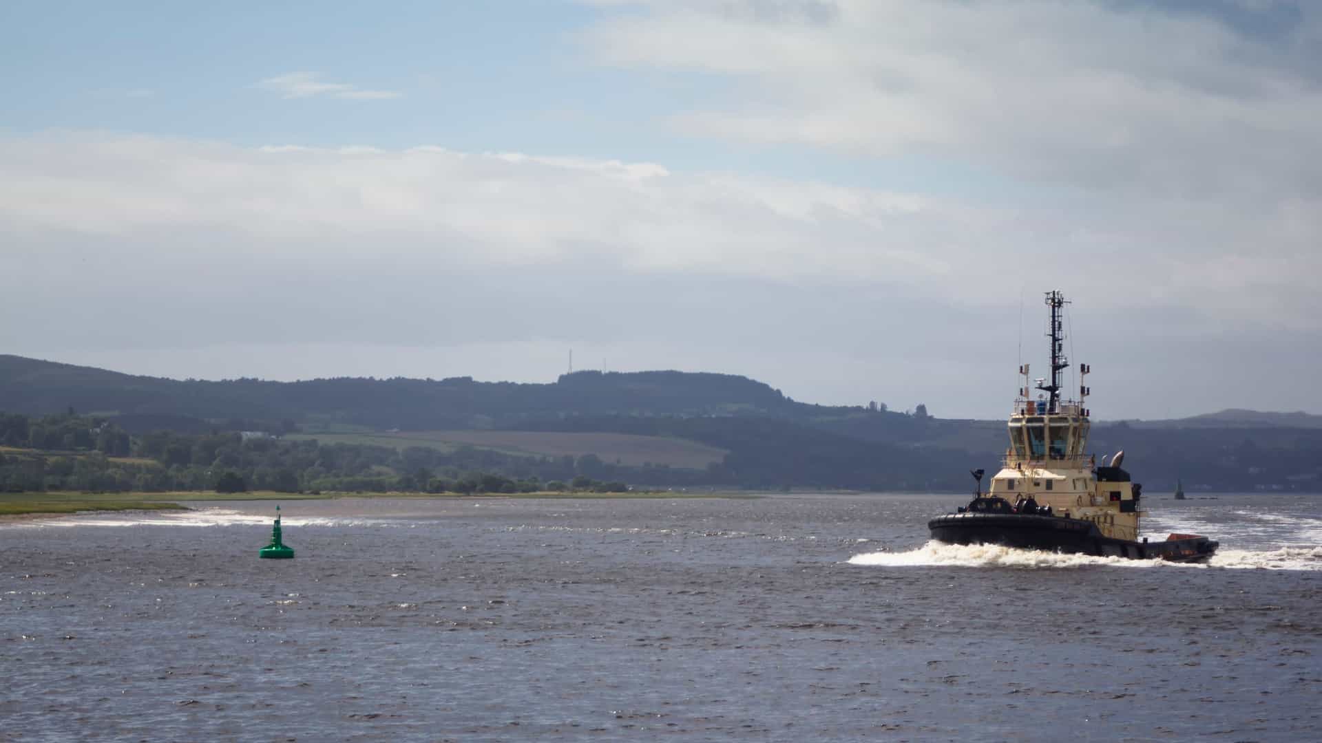 A brightly colored, powerful tugboat, predominantly red and blue, moves across the choppy waters of the River Clyde in Scotland. The tugboat is heading toward the right side of the frame with the industrial docks and structures of the Greenock port visible in the background, all set against a muted, cloudy gray sky.