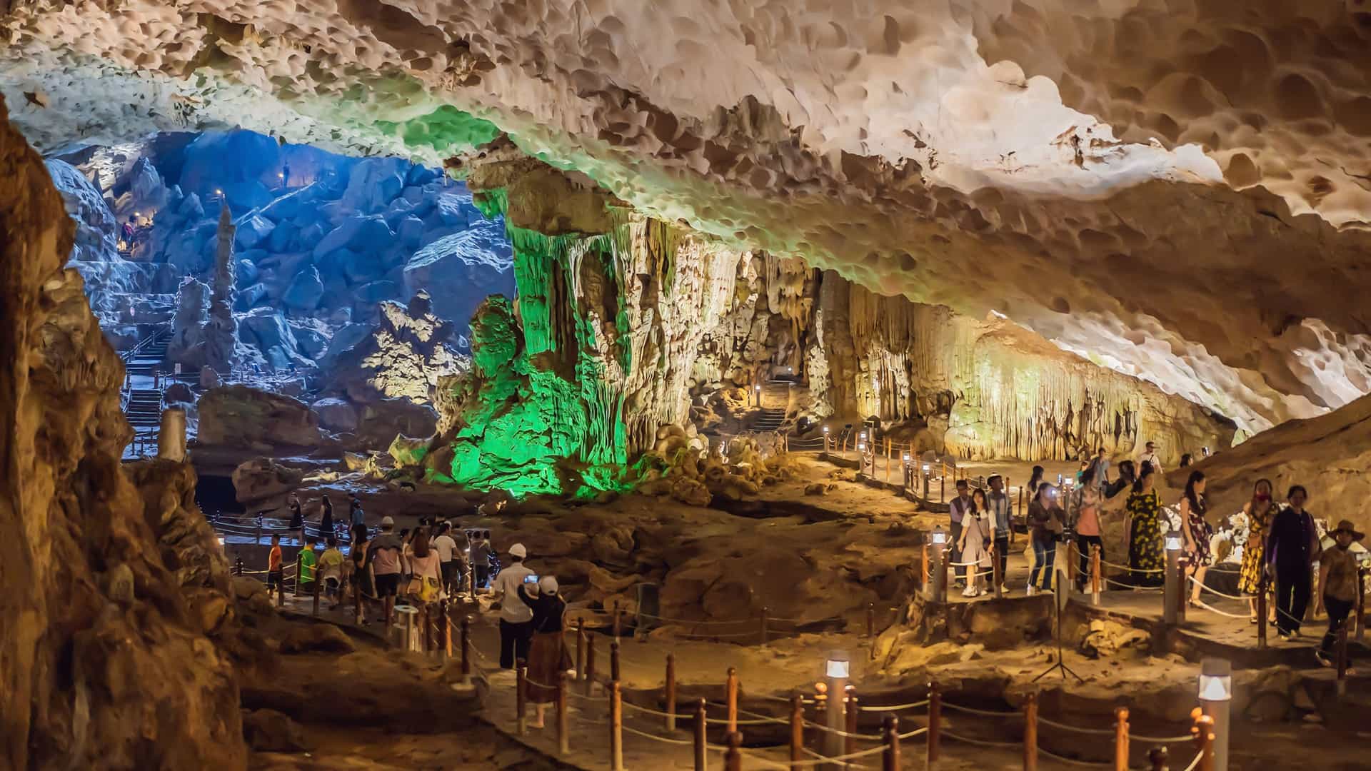  A view from a high vantage point inside the Sung Sot Grotto (Surprise Cave) in Ha Long Bay, Vietnam. The immense cave chamber is illuminated with colorful, artificial lights that highlight the towering stalactites and stalagmites. A paved pathway winds through the massive space, leading visitors deeper into the cave's dramatic and otherworldly interior.