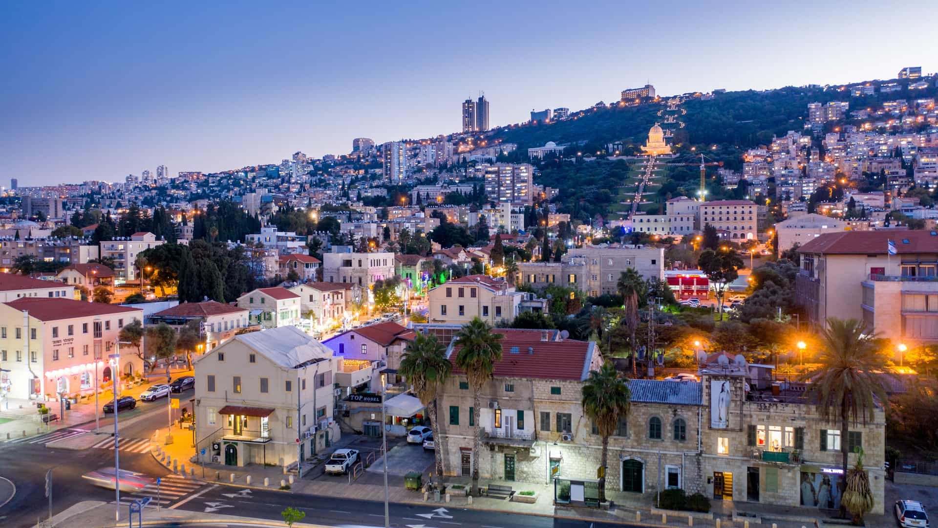 A stunning night aerial panorama of the illuminated German Colony (Moshavá Germanit) district in Haifa, Israel. The view looks down the brightly lit main street, with the port and harbor visible in the foreground, and the spectacular, terraced Baháʼí Gardens and Shrine of the Báb rising up the mountain in the background, their fountains and tiers glowing under artificial light.