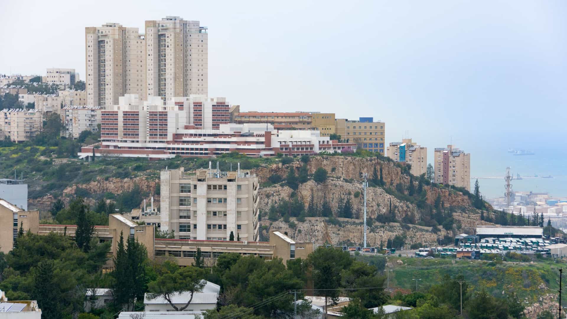 A wide, bird's-eye view of the Technion – Israel Institute of Technology campus in Haifa, Israel. The large complex of buildings and green spaces is set against the forested Carmel Mountains in the background, with a panoramic vista of Haifa Bay and the Mediterranean Sea visible in the distance on the right.