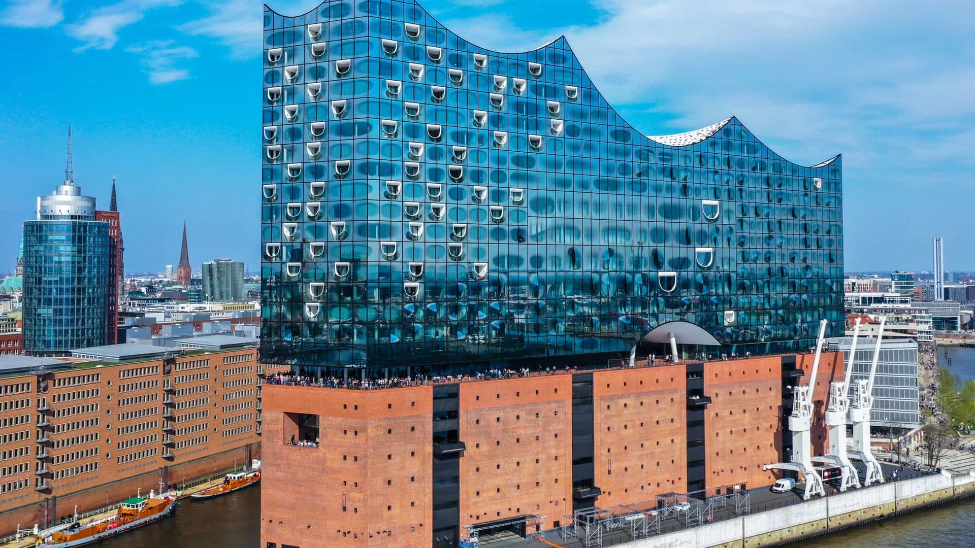 -A stunning architectural view of the Elbphilharmonie (often nicknamed 'Elphi') concert hall in Hamburg, Germany. The building features a wavy, tent-like glass structure resting atop the historic red brick foundation of the former Kaispeicher A warehouse, creating a dramatic contrast against the harbor waterfront.