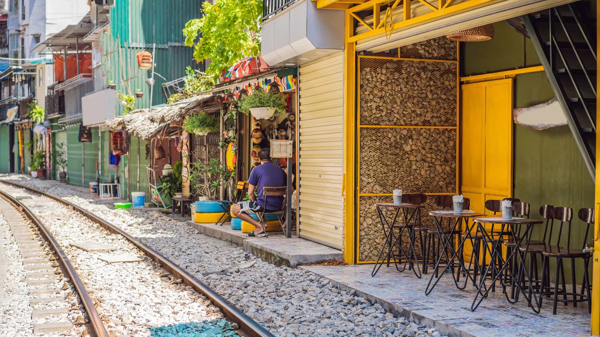  A photograph of Hanoi's "Train Street," a narrow railroad track that runs directly through a residential alley. The view is from a low angle, showing the train tracks in the center, with colorful, old houses and small businesses lining the tracks on both sides. People are visible sitting at small cafes or standing close to the buildings, and a few lanterns and other decorations are hanging overhead.