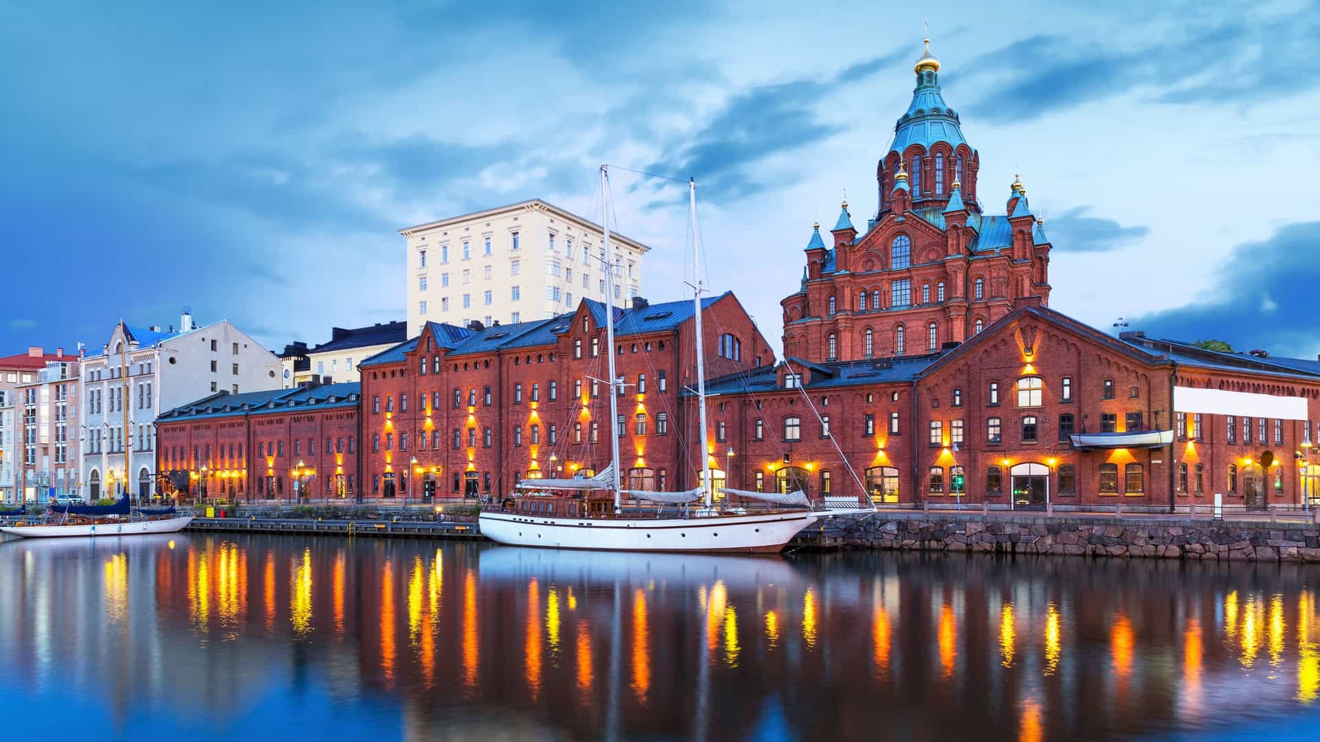  A photograph of the Uspenski Orthodox Cathedral in Helsinki, Finland, at dusk. The cathedral's red brick facade and a number of its golden onion domes are illuminated against a dark evening sky. The church stands on a rocky hill overlooking the city, and the photograph captures its prominent, historic architecture.