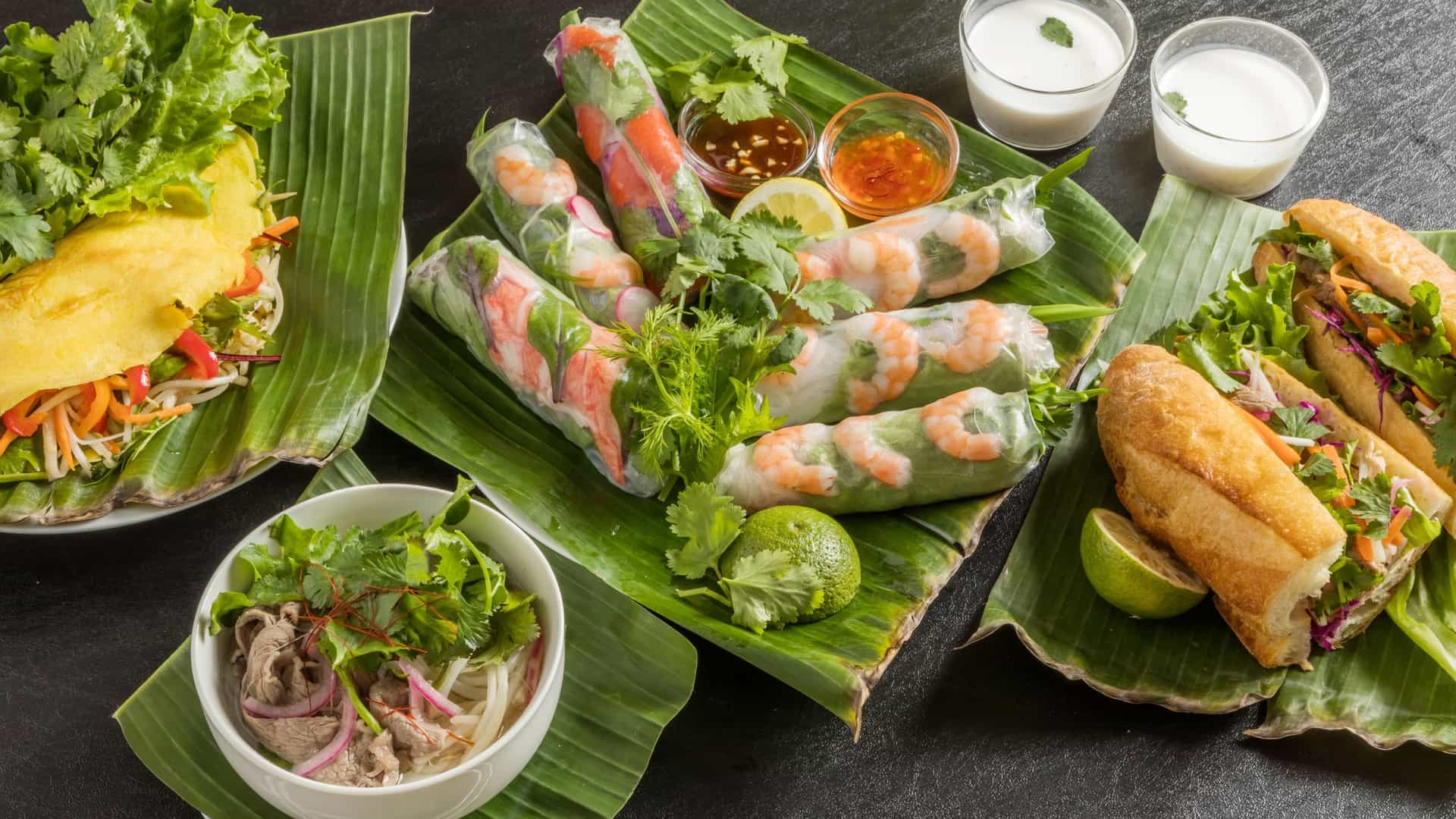  A photograph of two classic Vietnamese dishes. On the left is a bowl of Pho, a rice noodle soup with a rich broth, garnished with fresh herbs, bean sprouts, and lime wedges. On the right is a Banh mi sandwich, a baguette filled with various ingredients, including pickled vegetables, herbs, and a spread of pâté. The food is arranged on a table, highlighting the vibrant colors and textures of the dishes.