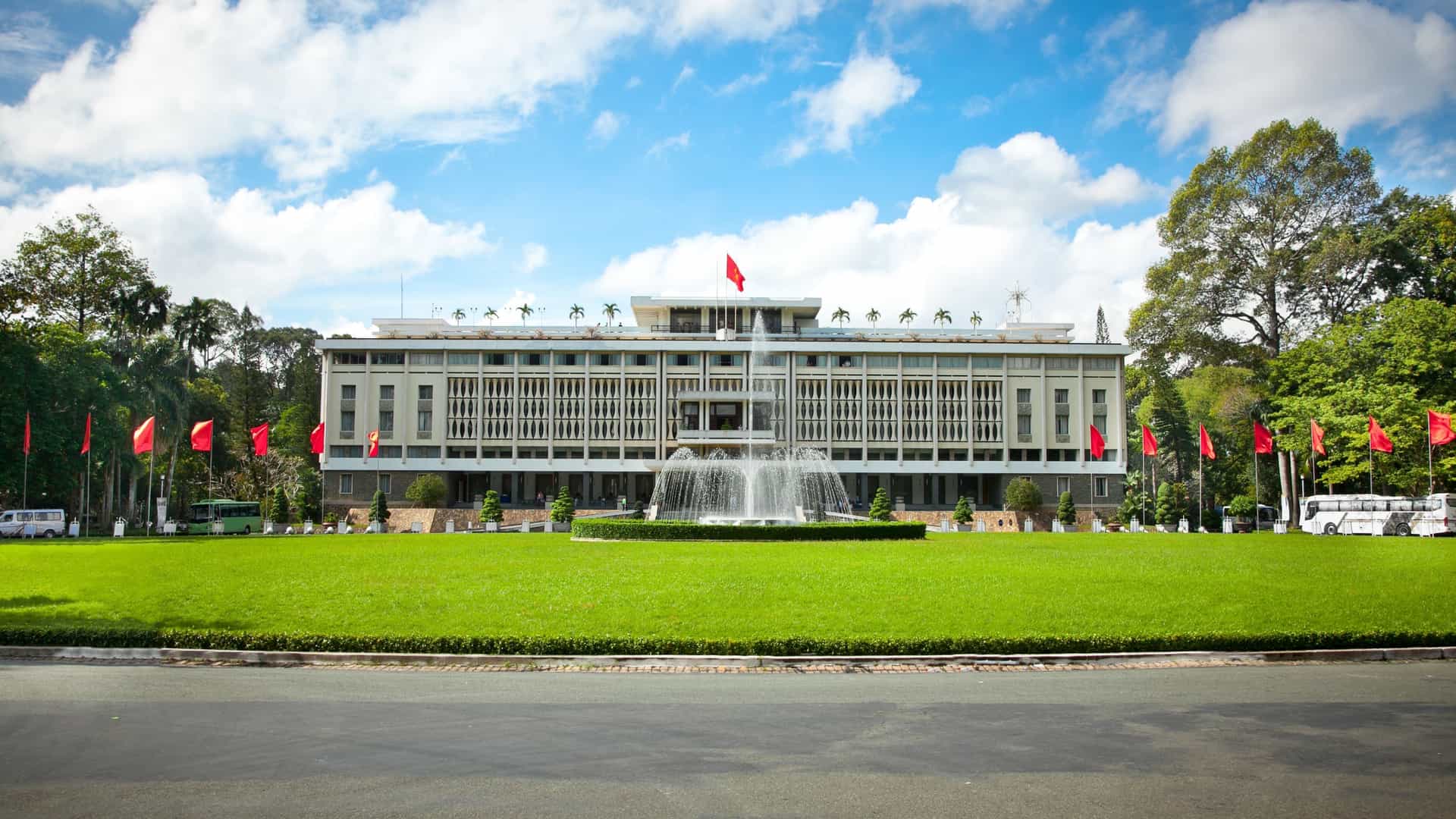  A daytime photograph of the Reunification Palace (also known as Independence Palace) in Ho Chi Minh City, Vietnam. The image shows the modernist, 1960s era building with its striking geometric architecture. The palace is surrounded by lush green lawns and tropical trees. The wide, open space in front of the building highlights its monumental size and historical significance.
