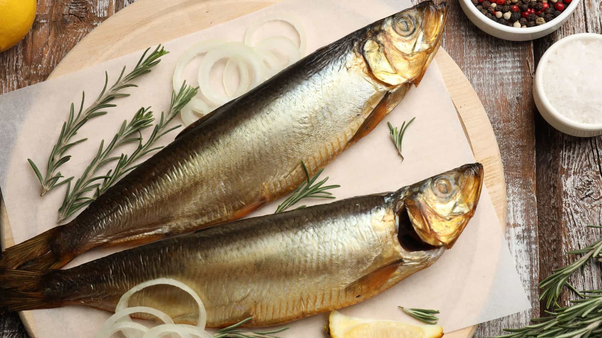 A close-up, top-down (flat lay) view of several pieces of whole or filleted smoked herrings arranged on a rustic wooden table or board, accompanied by various spices, peppercorns, and herbs, highlighting a savory and rustic food scene.