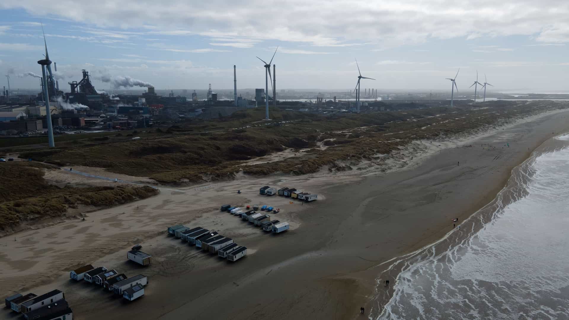 An aerial photograph capturing the modern landscape of IJmuiden, North Holland, Netherlands. The image shows a mix of coastal buildings, industrial structures, and large, contemporary wind turbines (windmills) on the land or just offshore, symbolizing the town's connection to industry and renewable energy.