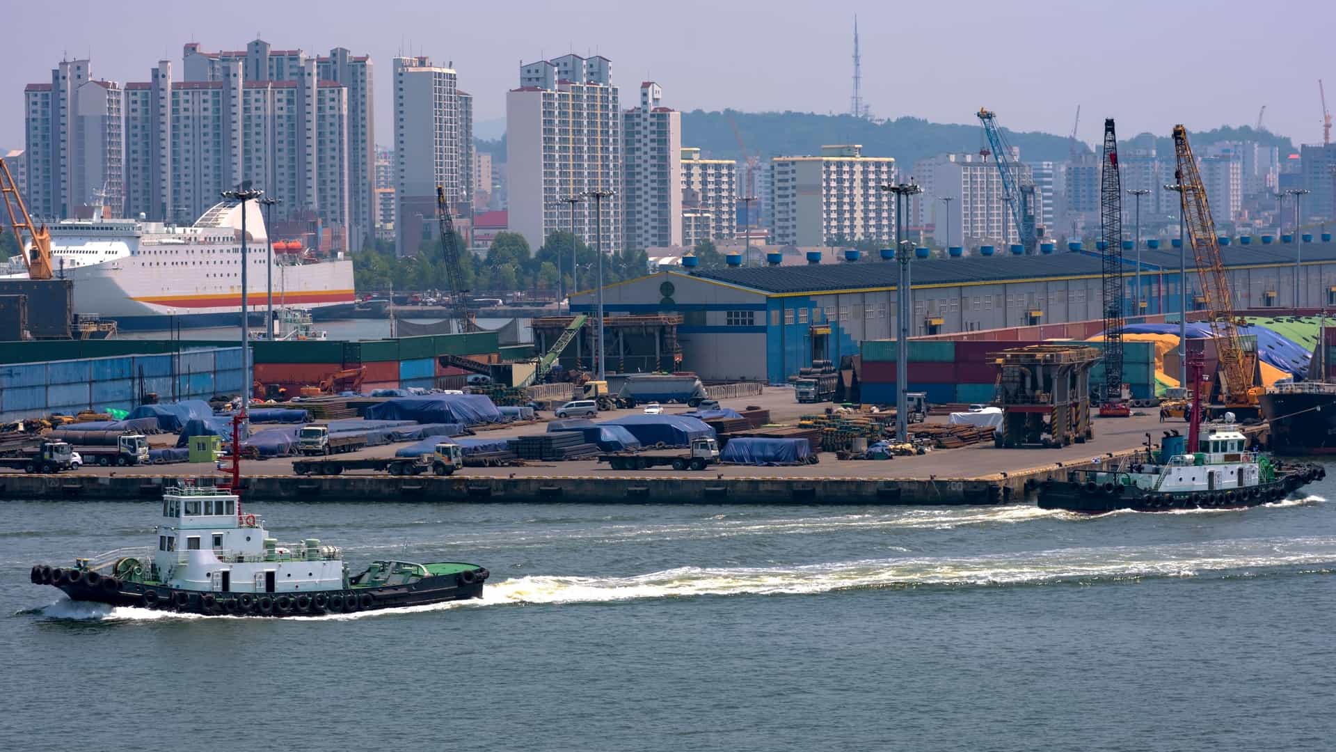 A view of the busy Incheon Port in South Korea, showing the active harbor with two white and green tugboats moving across the foreground. In the background is the industrial port with shipping containers, cranes, a large passenger ferry, and the dense, modern high-rise skyline of the city, which is part of the larger Incheon Open Port Area.