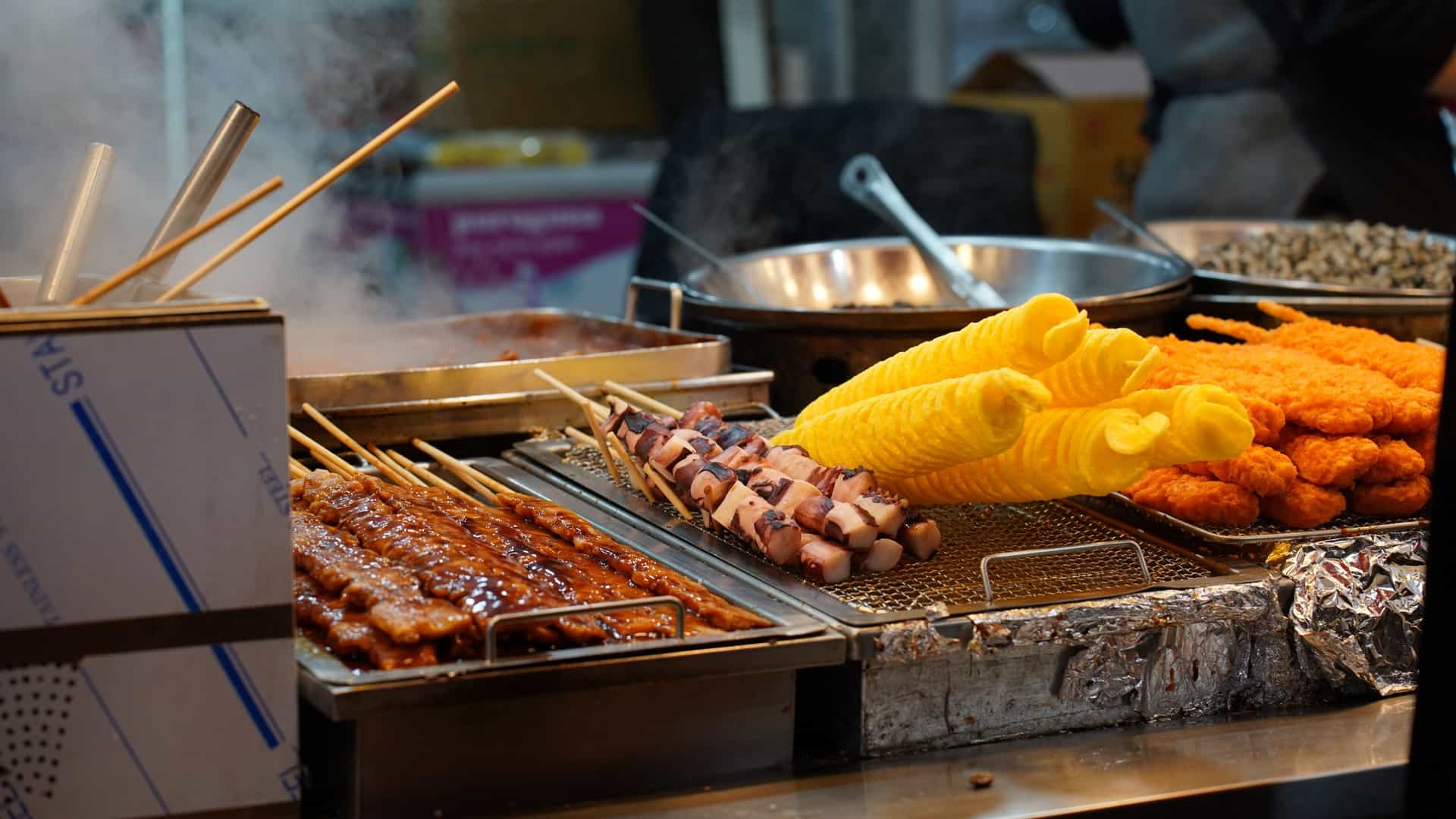 A close-up of a vibrant Korean street food stall, representative of the food found at places like Sinpo Market. The stall showcases a variety of popular grilled and fried snacks, including glazed meat skewers, skewers of octopus or sausage pieces, and large, bright yellow, battered items (like tornado potatoes or corn), all being kept hot on the metal griddle.