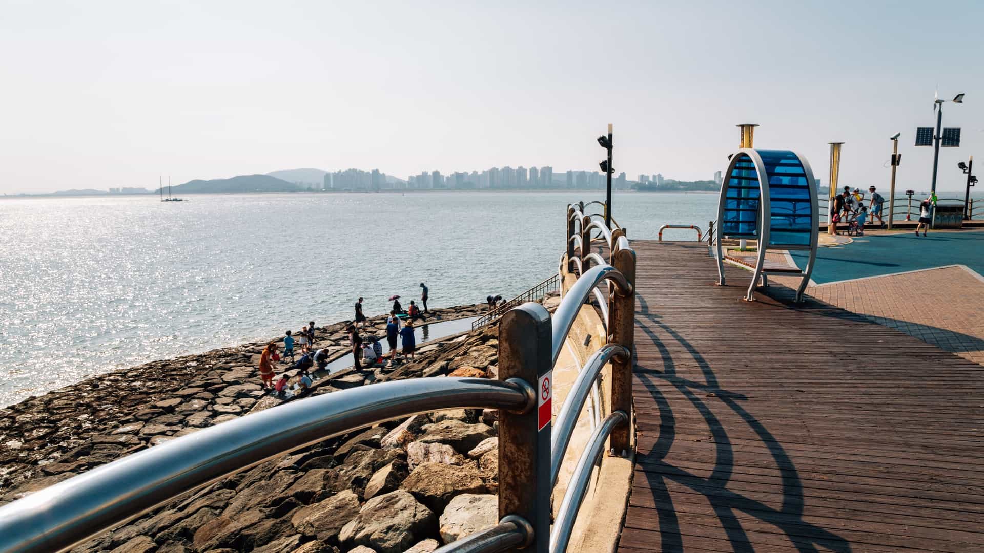 A view of the Wolmido Island Park Seaside Road in Incheon, South Korea. The image likely features the coastal promenade or street that runs along the water, often showing the sea, part of the Wolmi Theme Park (like the Ferris wheel), and the modern city skyline of Incheon in the distance.