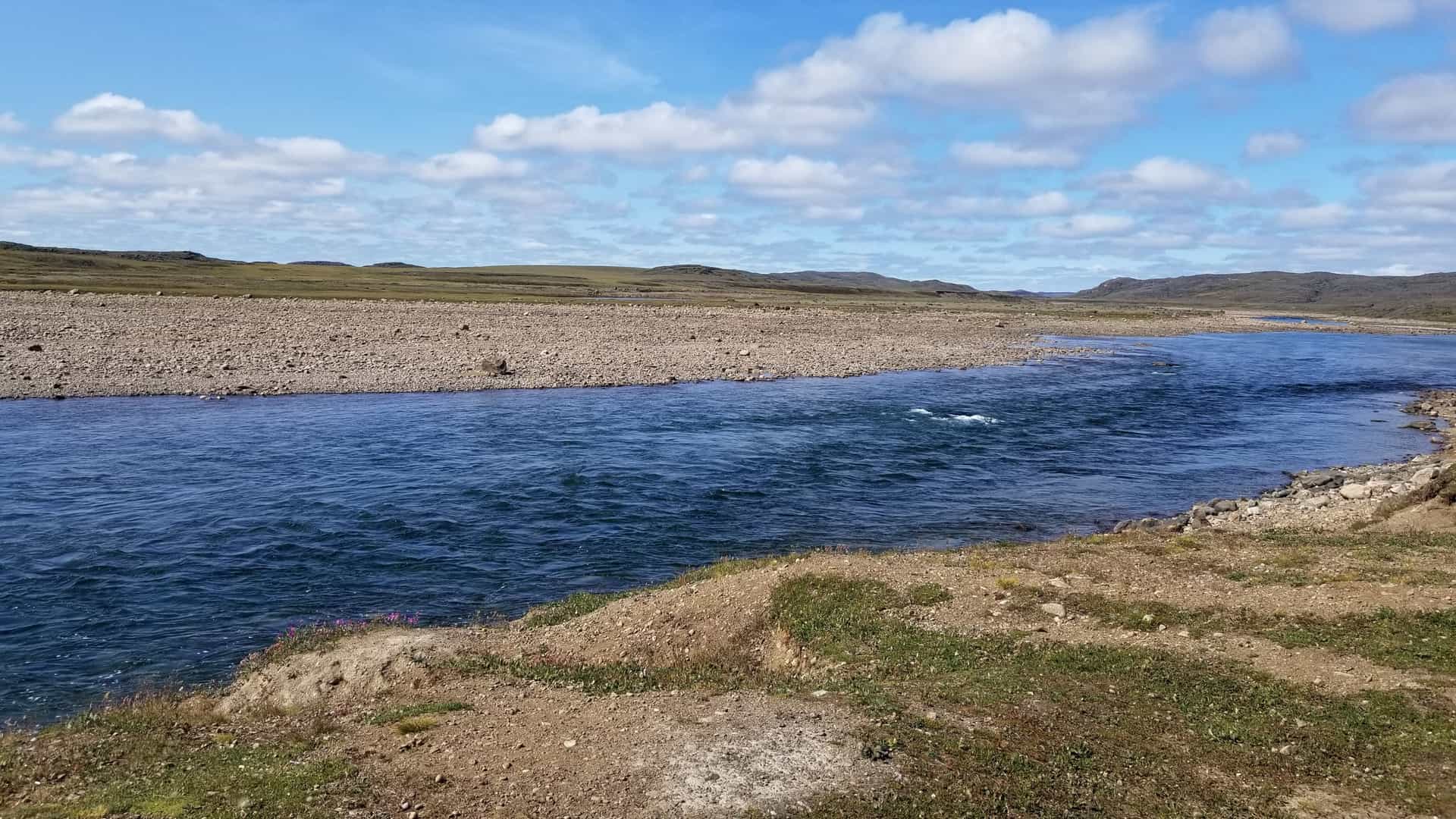 A wide-angle landscape view of the Sylvia Grinnell River flowing through the Sylvia Grinnell Territorial Park near Iqaluit, Nunavut. The deep blue river dominates the foreground and middle ground, bordered by a wide, rocky, treeless expanse of Arctic tundra under a bright blue sky scattered with white clouds.
