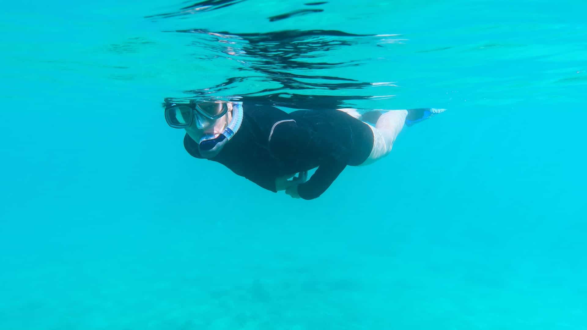 An underwater photograph of a girl wearing a mask and snorkel while actively snorkeling in the waters around Isla Baltra in the Galapagos Islands. The clear, blue ocean water is visible, along with the rocky seabed below and sunlight filtering down from the surface.