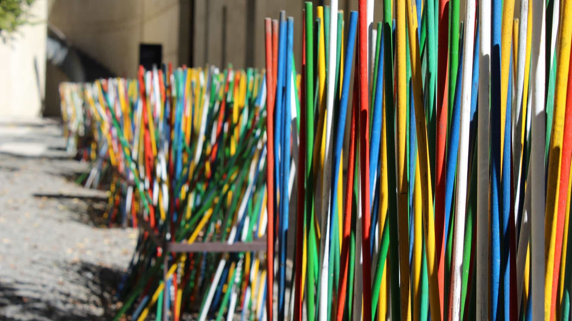 A vibrant abstract art installation at the Apartheid Museum in Johannesburg, South Africa. The piece features numerous closely placed, narrow wooden slats painted in a full spectrum of colors—red, orange, yellow, green, and blue—creating a textured, rainbow-like wall that symbolizes the contrast of post-apartheid life against the museum's black-and-white theme.