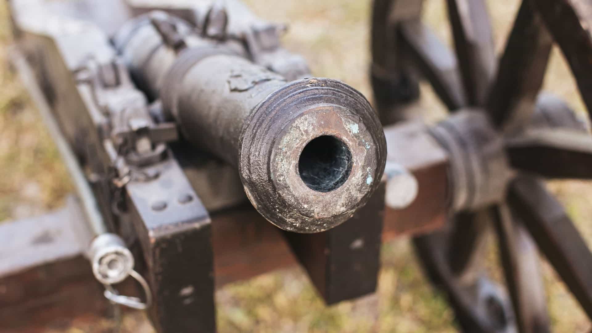 A detailed, focused, close-up view of the muzzle and barrel of an antique, dark metal artillery cannon, situated on a wooden carriage with a large spoked wheel visible in the background. The cannon is displayed on a grassy area, representing an outdoor exhibit at the South African National Museum of Military History in Johannesburg.