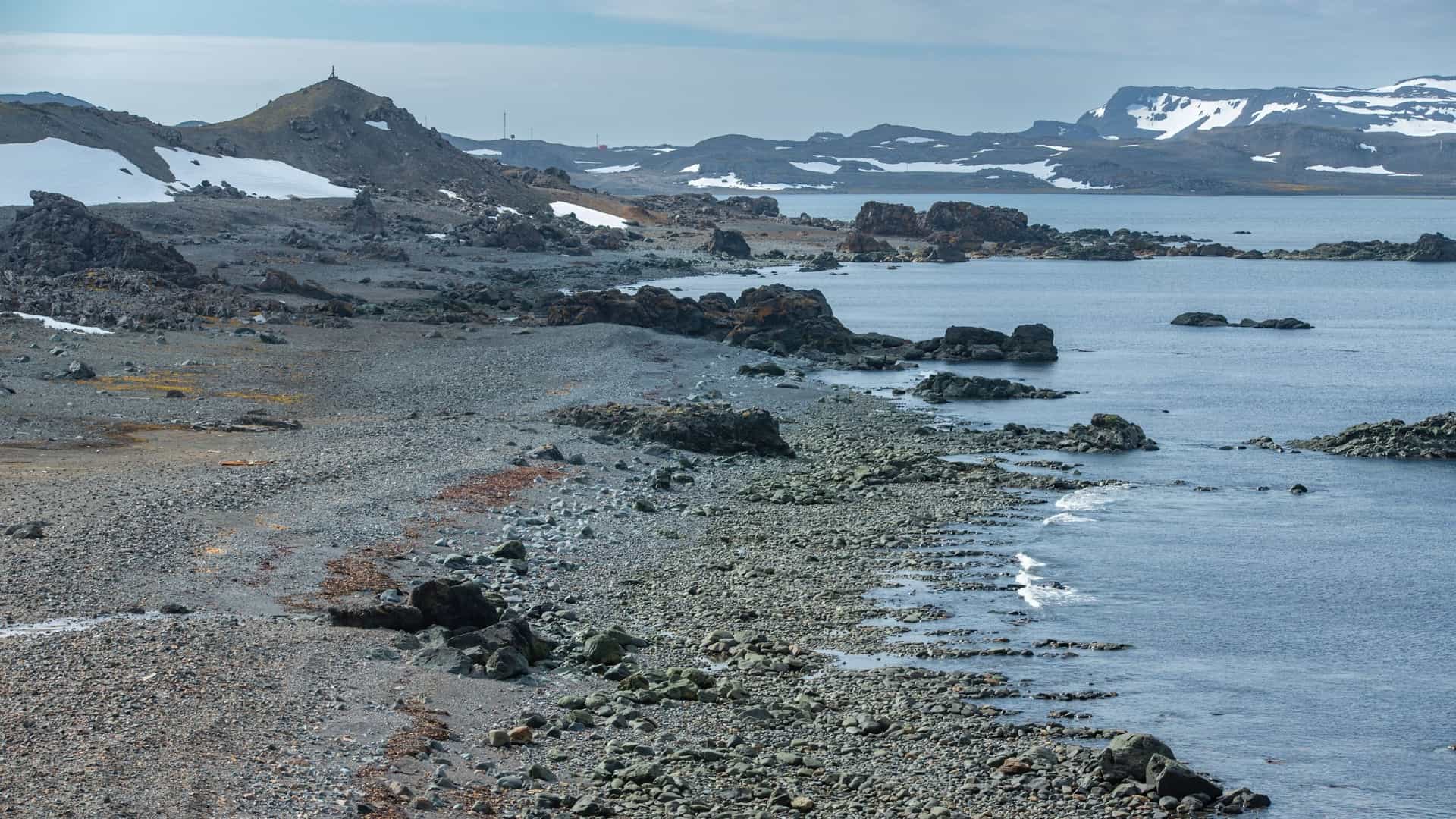 A wide, rugged landscape shot showing a desolate Antarctic coastline featuring a wide, dark gravel and rock beach meeting the calm, gray-blue ocean water. The rocky, volcanic terrain slopes up to barren hills where patches of snow remain on the distant peaks and on the dark, rocky ground—a stunning, if frigid, environment to Enjoy a Picnic with a View.