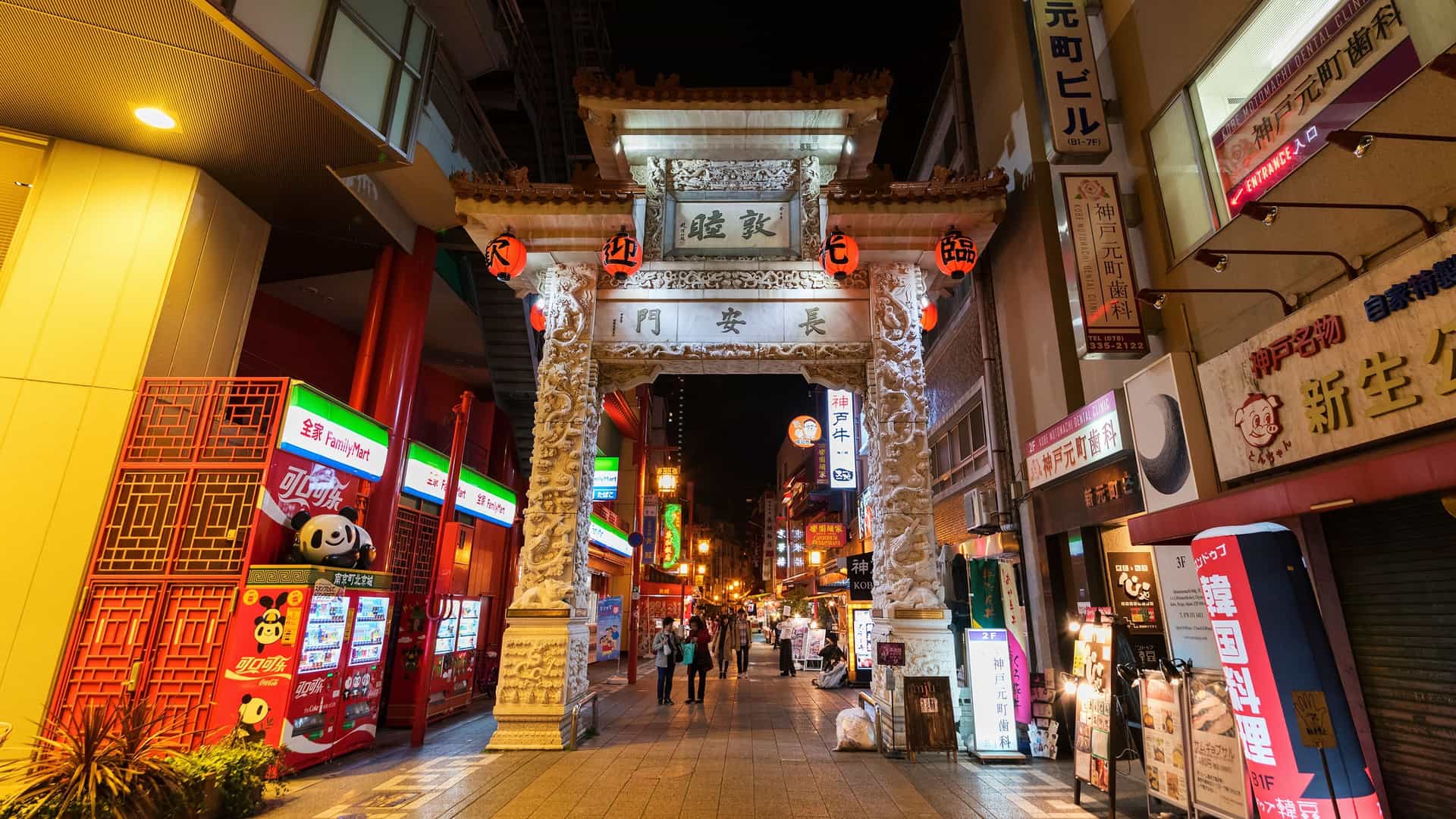 A vibrant street scene in Kobe's Nankinmachi Chinatown, with a large, ornate red and yellow Chinese gate (likely the Choanmon Gate) in the background. The street is bustling with people and is lined with food stalls and shops adorned with numerous red Chinese lanterns and colorful banners, showcasing the lively atmosphere of the district.