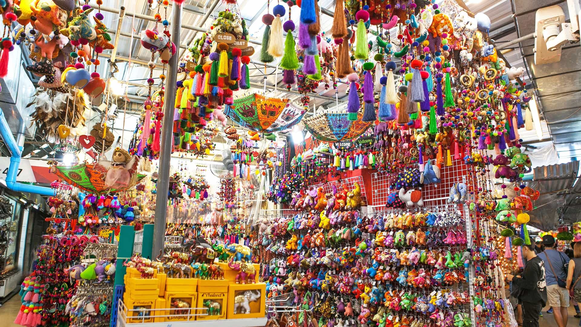 A colorful display of Thai-style souvenirs for sale at a stall in the Chatuchak Market in Bangkok, which is described as the largest market in Thailand. The image features a variety of small, decorative objects and handicrafts, likely including textiles, carvings, or trinkets, arranged for browsing.