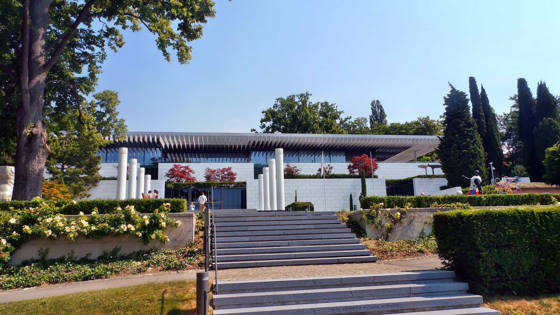 A wide exterior view of The Olympic Museum in Lausanne, Switzerland. The image showcases the modern, white, multi-level building and its surrounding grounds, which include an outdoor staircase lined with decorative posts and an expansive park area with mature trees and landscaping. The scene is set under a bright sky.