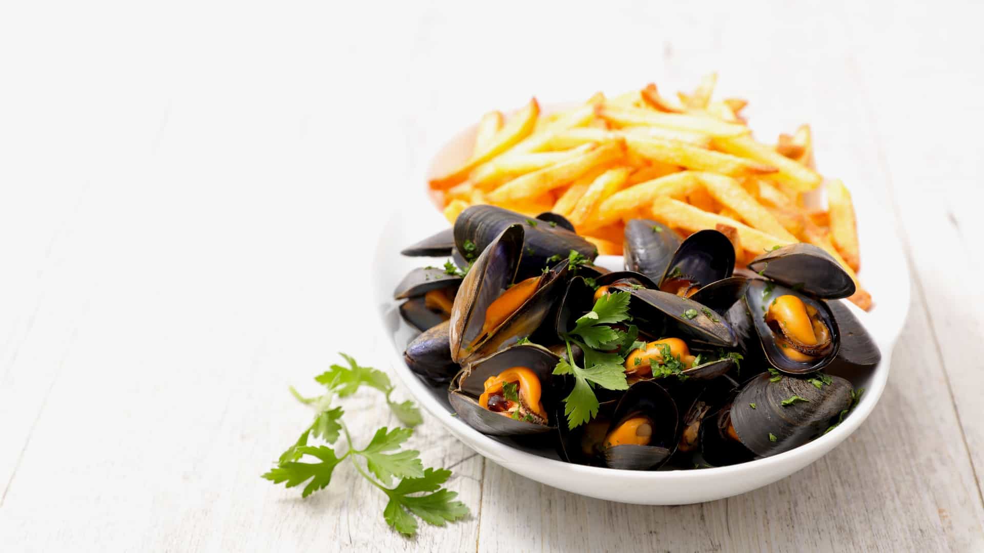 A close-up of Moules-frites, the traditional Belgian and French dish. A large, deep metal pot of fresh steamed mussels (moules) sits on a table, often alongside a basket or bowl of crispy golden French fries (frites). The dish is often served with a side of dipping sauce and a rustic, maritime background, emphasizing the culinary experience.