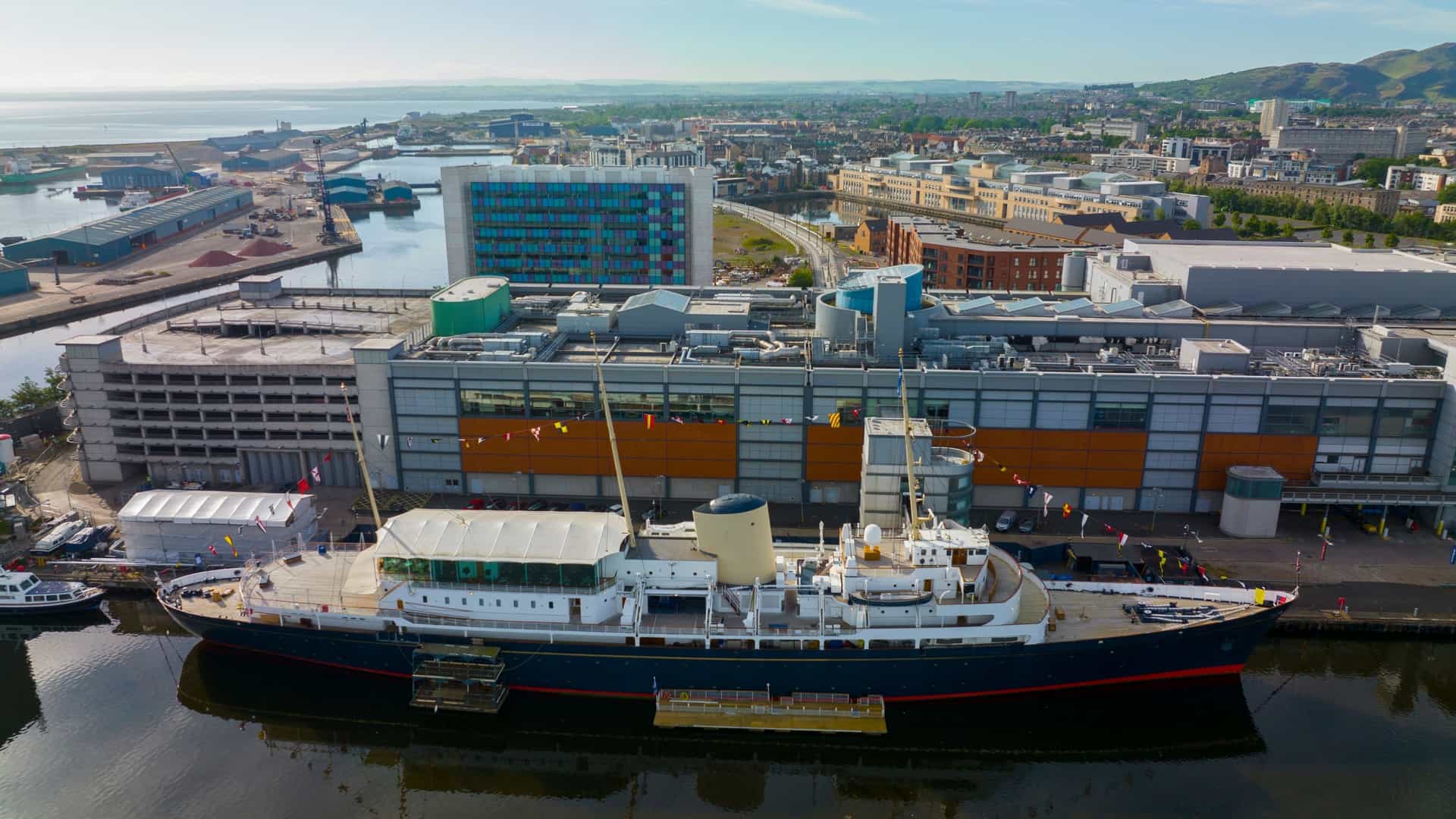 An aerial, top-down view of a beautifully restored antique steam yacht docked at a quay near a large port or Ocean Terminal. The image showcases the vessel's classic, elegant lines, wooden deck, and prominent smokestack, contrasting the historic ship with its modern surroundings and the blue of the water.