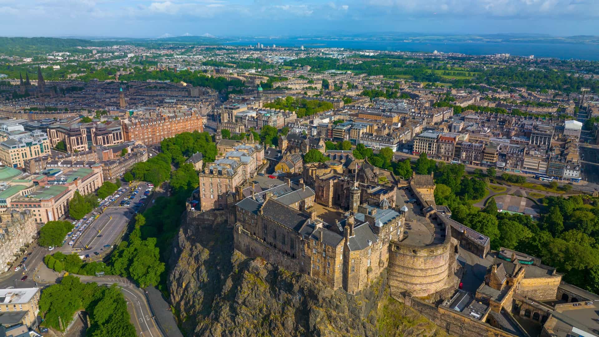 A dramatic, panoramic view of Edinburgh Castle, a historic fortress that stands proudly on the Castle Rock in Old Town Edinburgh, Scotland, with the stone walls dominating the steep volcanic crag. The castle is a prominent symbol of Scotland's history and a UNESCO World Heritage Site.