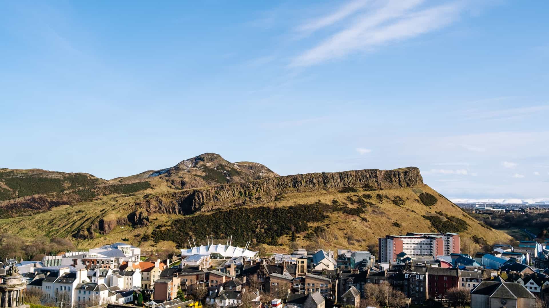 A breathtaking panoramic view of Edinburgh, Scotland, as seen from the summit of Arthur's Seat, an ancient extinct volcano. The cityscape stretches out below, featuring the historic buildings of the Old Town and the Firth of Forth in the distance, all set beneath a wide sky. Arthur's Seat is the highest point in Holyrood Park and a popular spot for hikers.