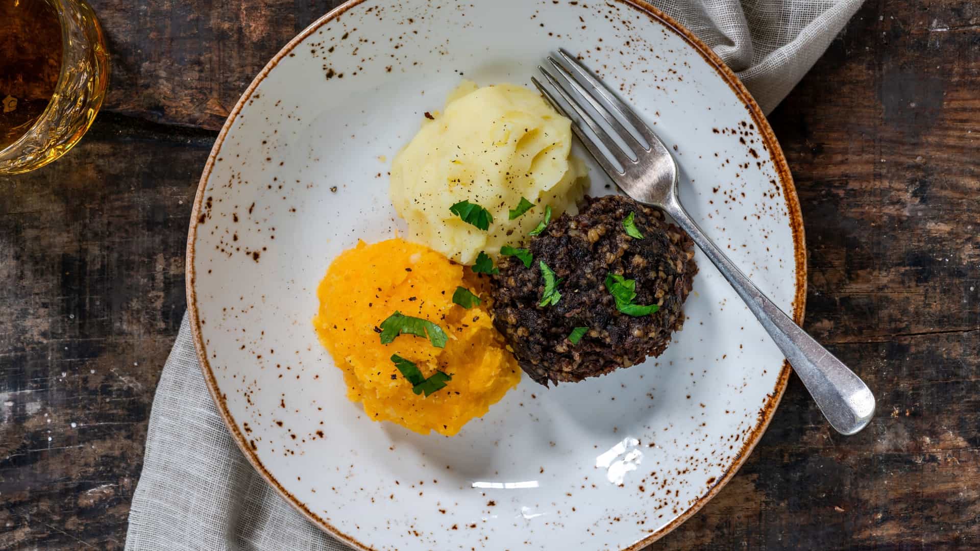 A close-up, top-down view of a serving of Haggis, Neeps, and Tatties, the traditional Scottish meal. The dish is presented with a mound of haggis (the savory pudding) surrounded by separate portions of mashed neeps (turnips or rutabaga) and mashed tatties (potatoes), typically served on a white plate.