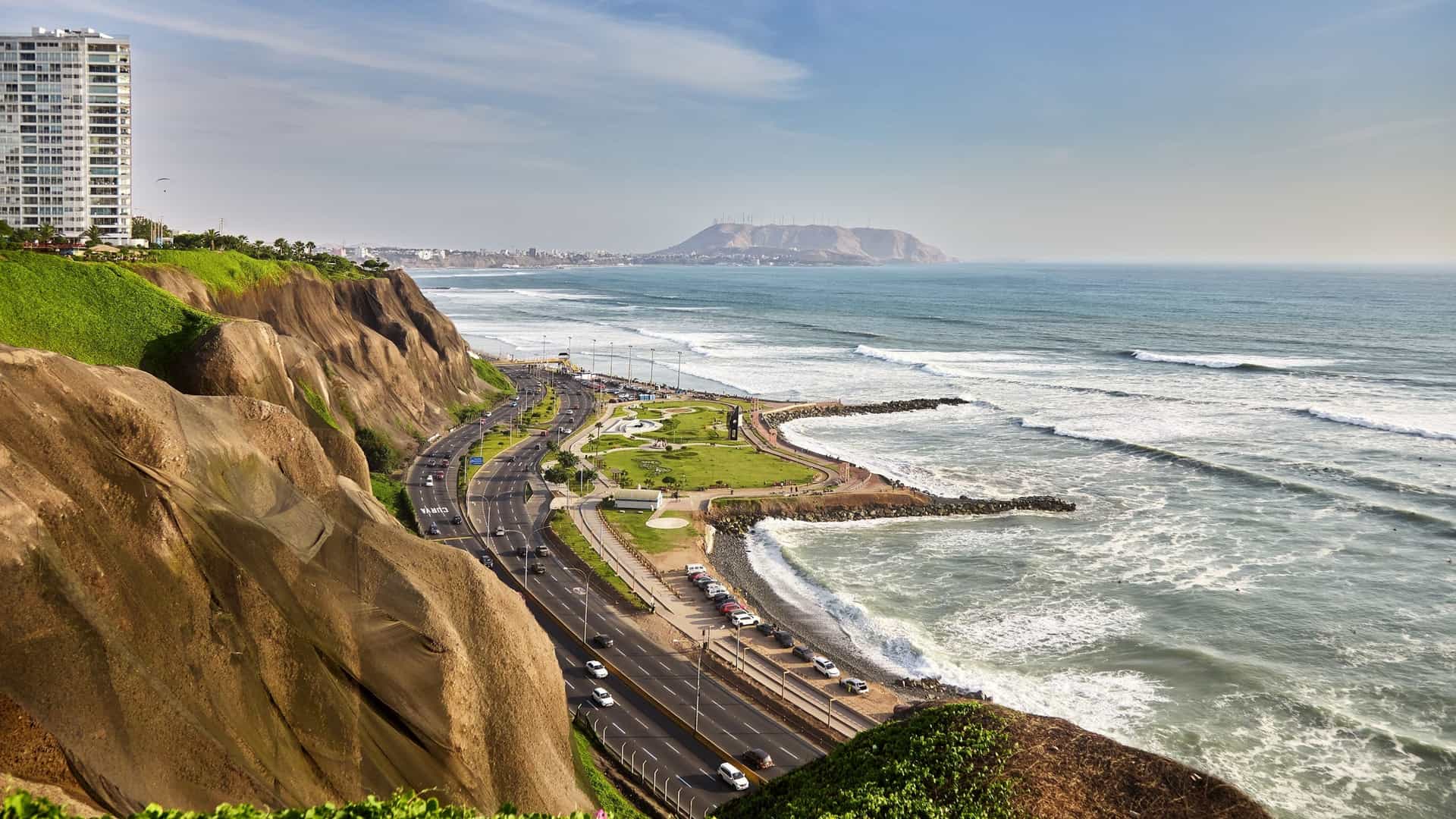 An aerial, panoramic view of the Miraflores district coastline in Lima, Peru, known as the Costa Verde (Green Coast). The image captures the dramatic, high cliffs covered in lush green parkland, with modern high-rise buildings perched along the top. Below the cliffs, the Pacific Ocean crashes onto a rocky beach, and the busy coastal highway is visible along the base. The scene is set under a bright sky, highlighting the dense urban area meeting the natural ocean-side bluffs.